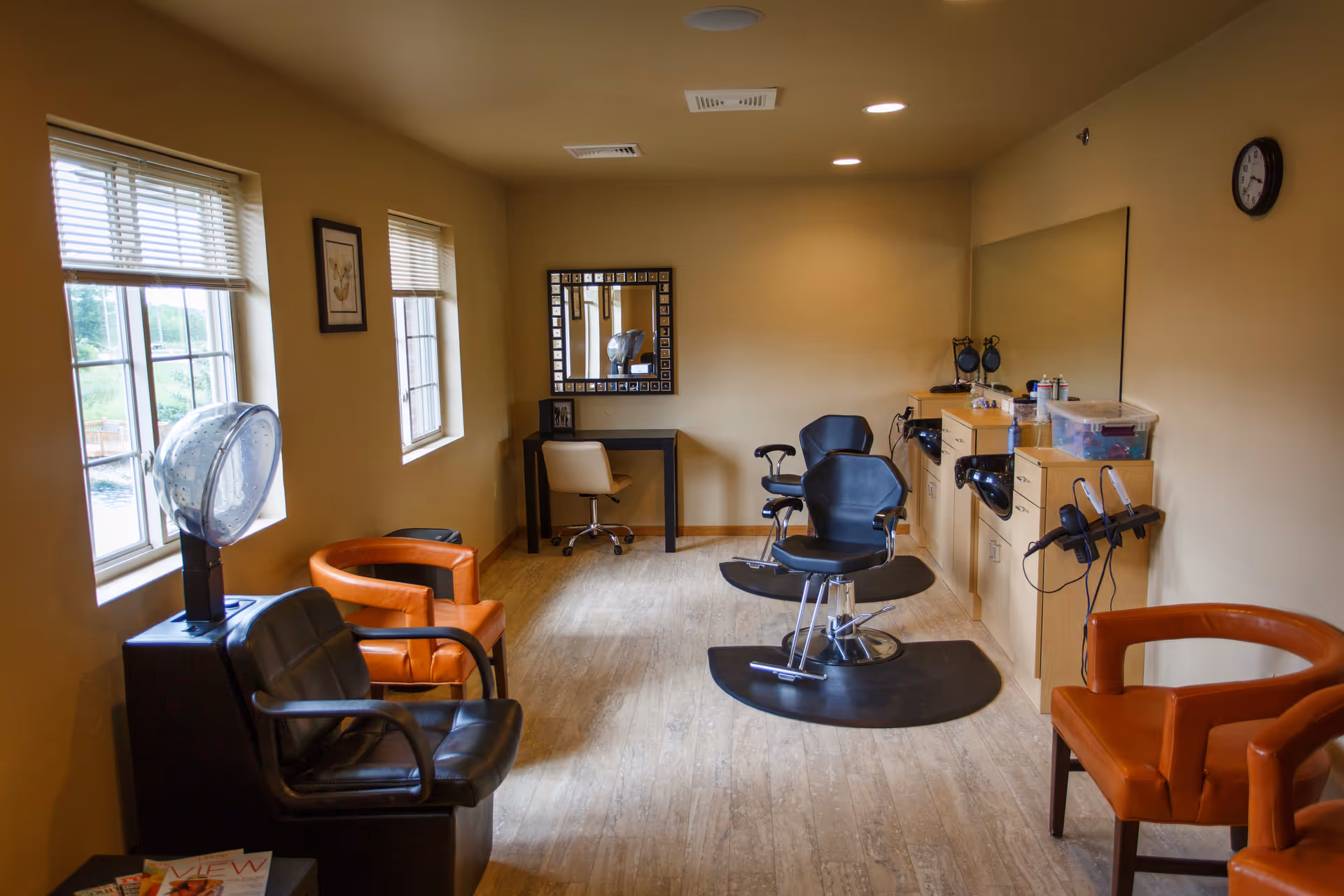 Interior view of a salon room in a senior living facility with two black salon chairs on mats, orange and black waiting chairs, a hair dryer, a large mirror, and a small desk with a chair. The room has beige walls, wood flooring, and two windows letting in natural light.
