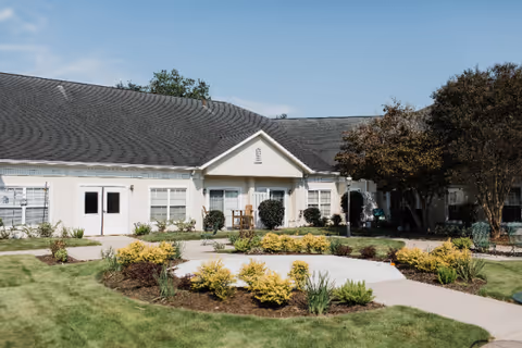 Exterior view of a single-story building with a dark shingled roof and white walls, surrounded by a well-maintained garden with green grass, shrubs, and small trees under a clear blue sky.