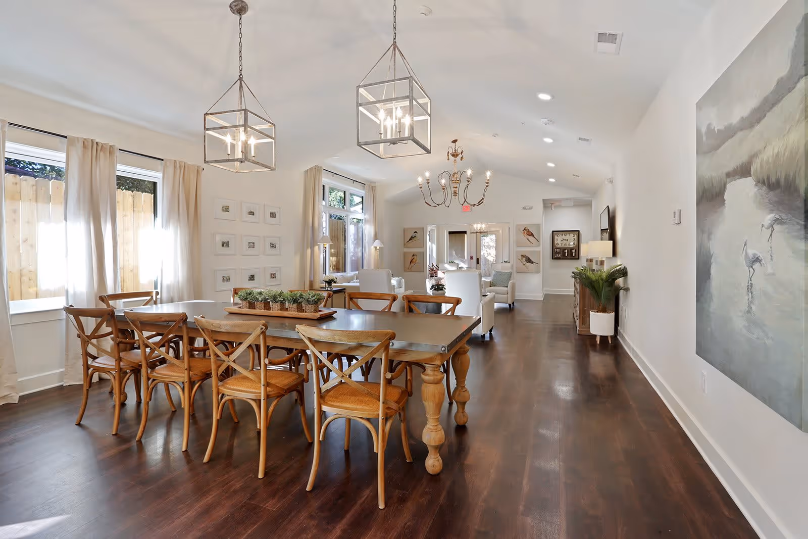 A bright and spacious dining and living area with a large wooden dining table surrounded by eight wooden chairs. The room features two modern pendant lights above the table, large windows with beige curtains, and a seating area with white armchairs and small tables in the background. The walls are decorated with framed pictures and a large painting of birds near a water body. The floor is dark wood, and the overall decor is light and airy.