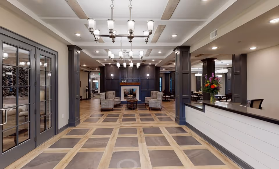 Spacious senior living facility interior with a checkered tile and wood floor pattern, dark wood columns and cabinetry, a fireplace with two armchairs and a small table in front, modern chandeliers hanging from the ceiling, a reception desk with a flower arrangement on the right, and glass double doors on the left.