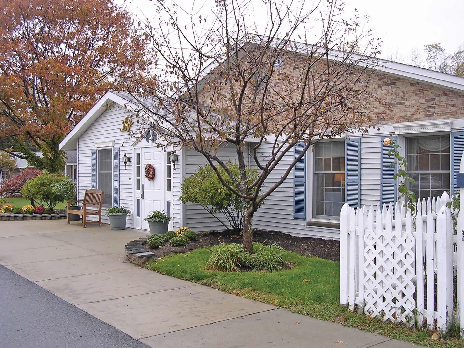 Exterior view of a single-story building with light gray siding and brick facade. There is a white door with a wreath, two windows with blue shutters, a white picket fence, a tree with sparse leaves, and some green shrubs and plants around the building. A wooden bench and potted plants are placed near the entrance. The sidewalk and part of the street are visible in the foreground.