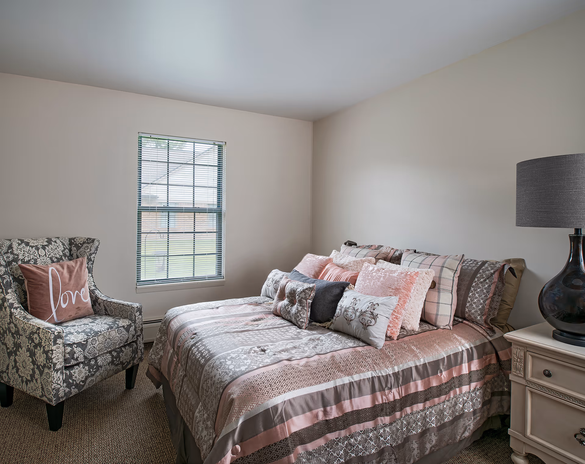 A cozy bedroom with a bed covered in patterned pink, gray, and white bedding and multiple decorative pillows. To the left of the bed is a floral upholstered armchair with a pink pillow that says 'love'. A window with blinds is on the wall behind the chair, and a nightstand with a dark lamp is on the right side of the bed.