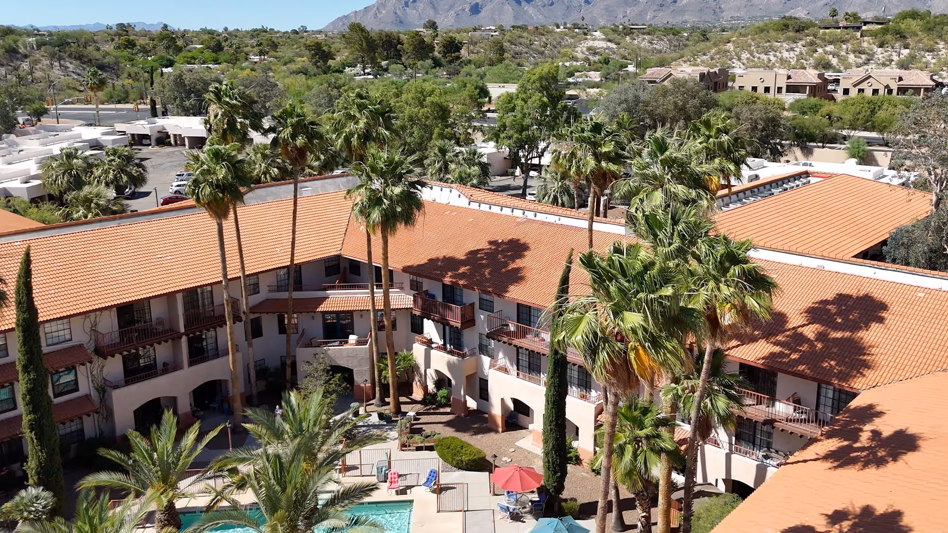 Aerial view of a senior living facility with a central courtyard featuring a swimming pool, surrounded by a three-story building with terracotta roof tiles and balconies. Tall palm trees and other greenery are scattered throughout the courtyard and surrounding area, with mountains visible in the background under a clear sky.