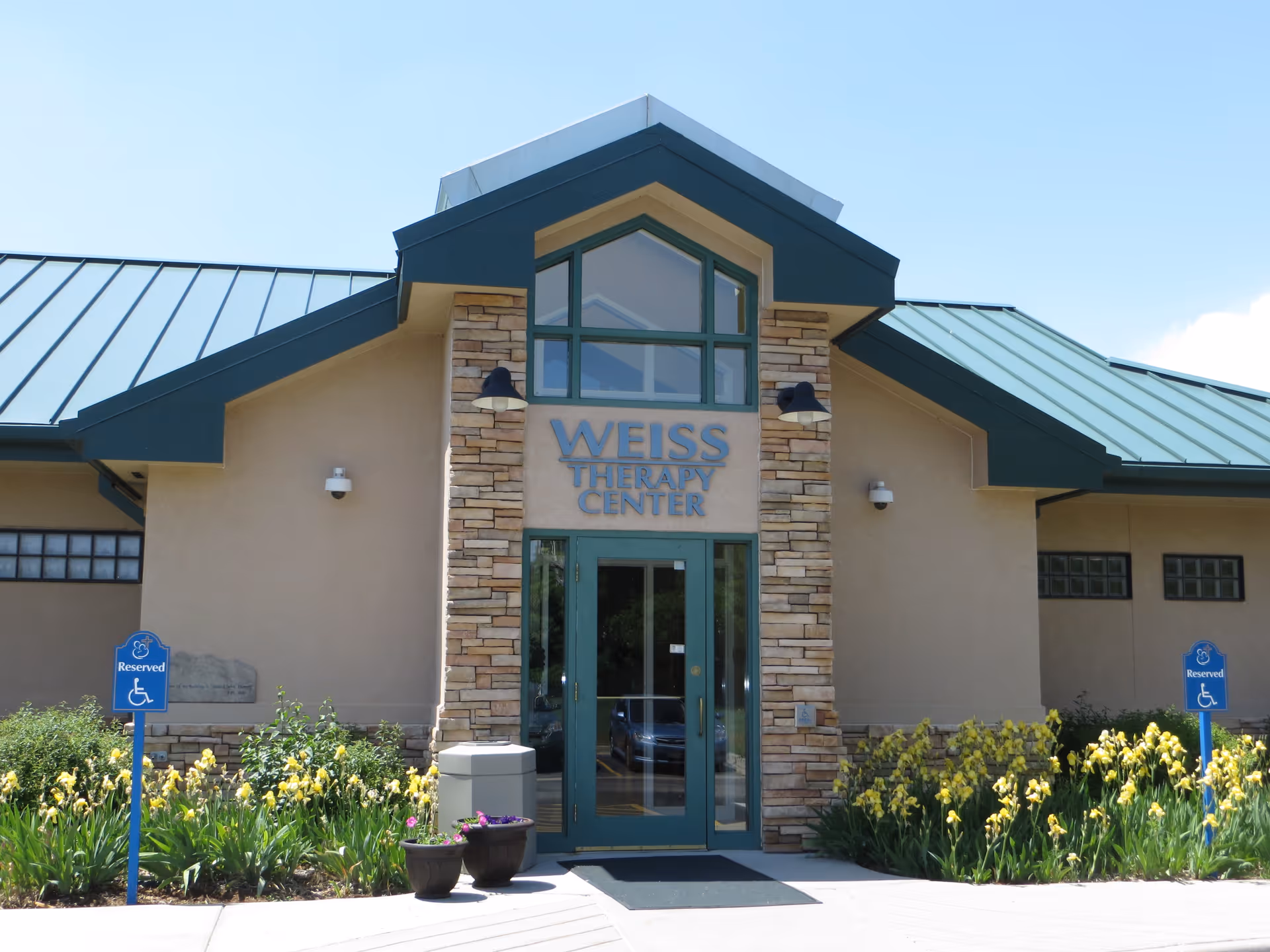 Exterior front entrance of the Weiss Therapy Center building with a green roof, stone accents around the door, and yellow flowers planted along the walkway. Two blue reserved handicap parking signs are visible on either side of the entrance.