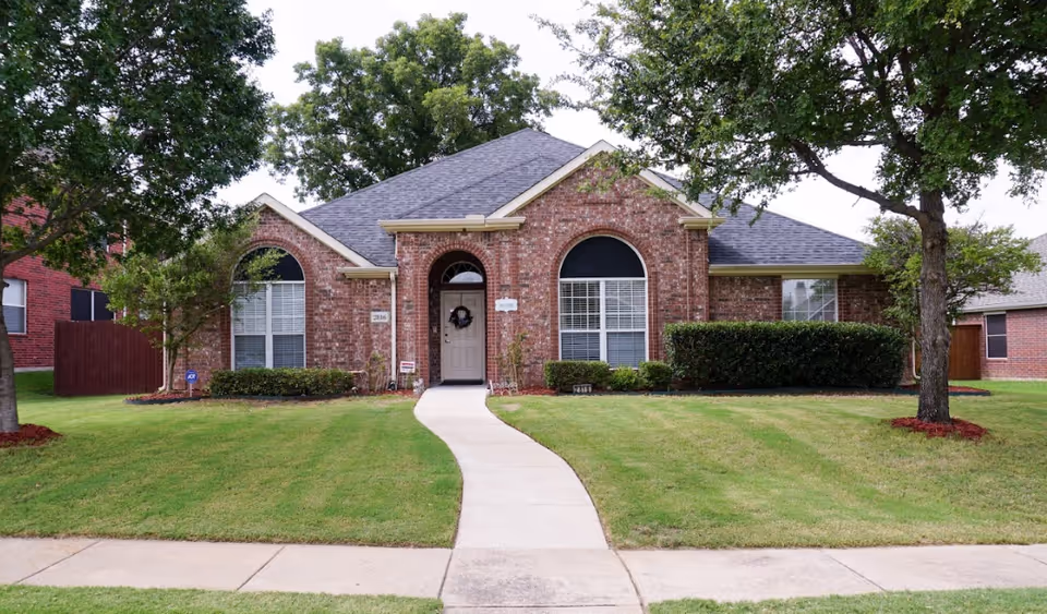 Front exterior view of a single-story brick house with a gray shingled roof, arched windows with black awnings, a central front door with a wreath, a curved concrete walkway leading to the entrance, green lawn, and trees on either side.