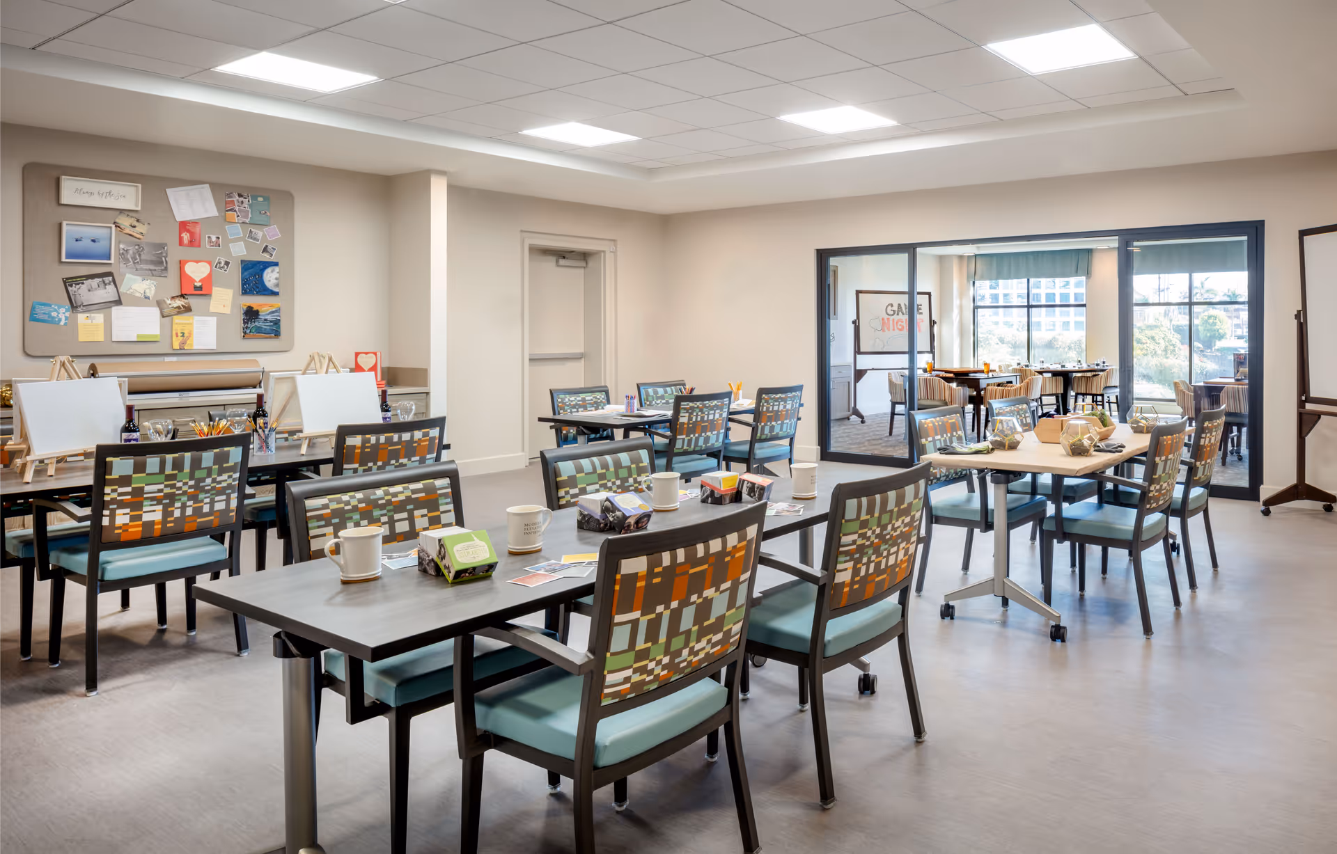 A bright, spacious activity and dining area in a senior living facility with multiple tables and chairs arranged neatly. The tables have cups, napkins, and some activity supplies like paintbrushes and easels. A bulletin board with various notes and pictures is visible on the wall. Large windows and glass doors lead to another room with more tables and chairs, and a sign that reads 'Game Night'.
