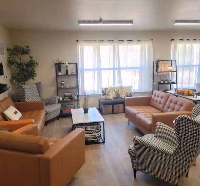 Bright communal living room with tan leather sofas, patterned armchairs, a coffee table, shelving, and large windows with sheer curtains.