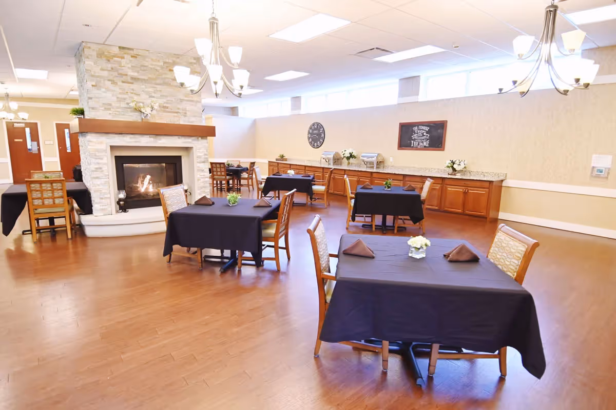 A dining room with several tables covered in black tablecloths, each set with folded brown napkins and small floral centerpieces. The room features a stone fireplace with a fire burning, wooden chairs, hardwood flooring, and a long counter with cabinets along the back wall. There are chandeliers hanging from the ceiling and a clock and framed sign on the wall.
