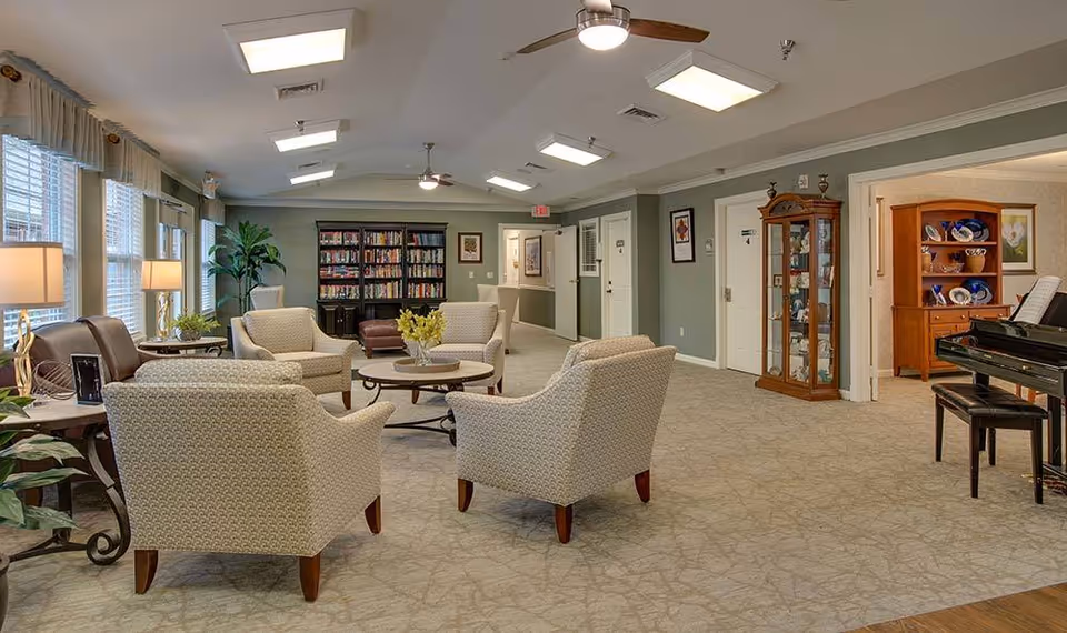 A spacious senior living common area with several cushioned armchairs arranged around a round coffee table. The room features large windows with curtains, a bookshelf filled with books, a piano with a bench, and display cabinets with decorative items. The walls are painted a soft green and the ceiling has recessed lighting and ceiling fans.