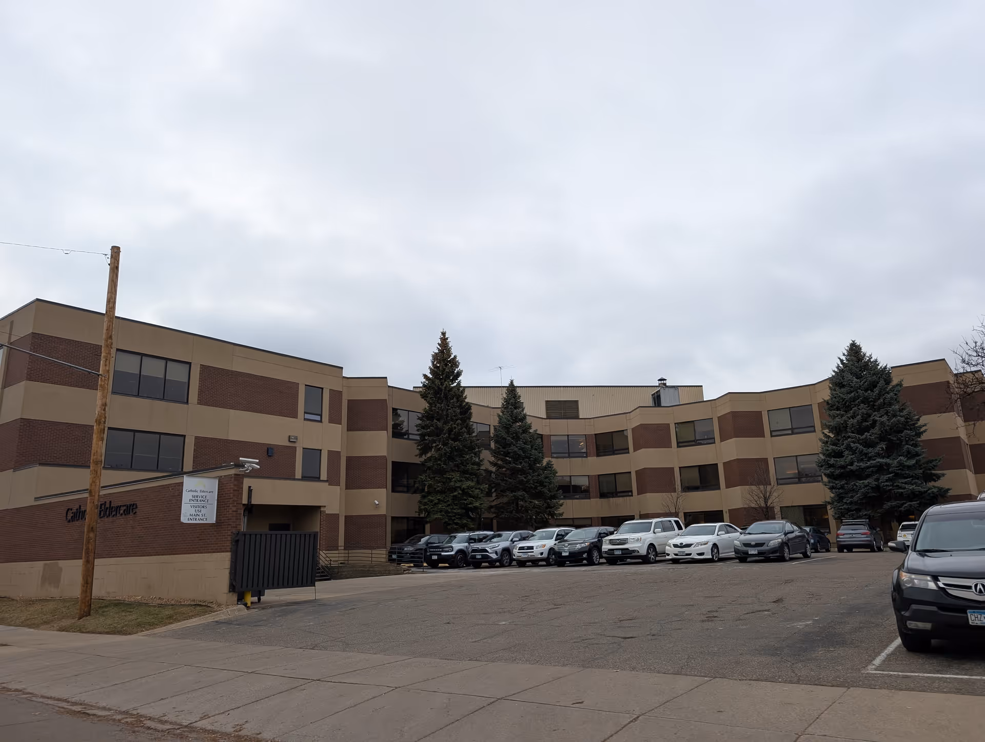 Exterior view of a three-story senior living facility building with a parking lot in front. Several cars are parked, and there are tall evergreen trees near the building. The sky is overcast.