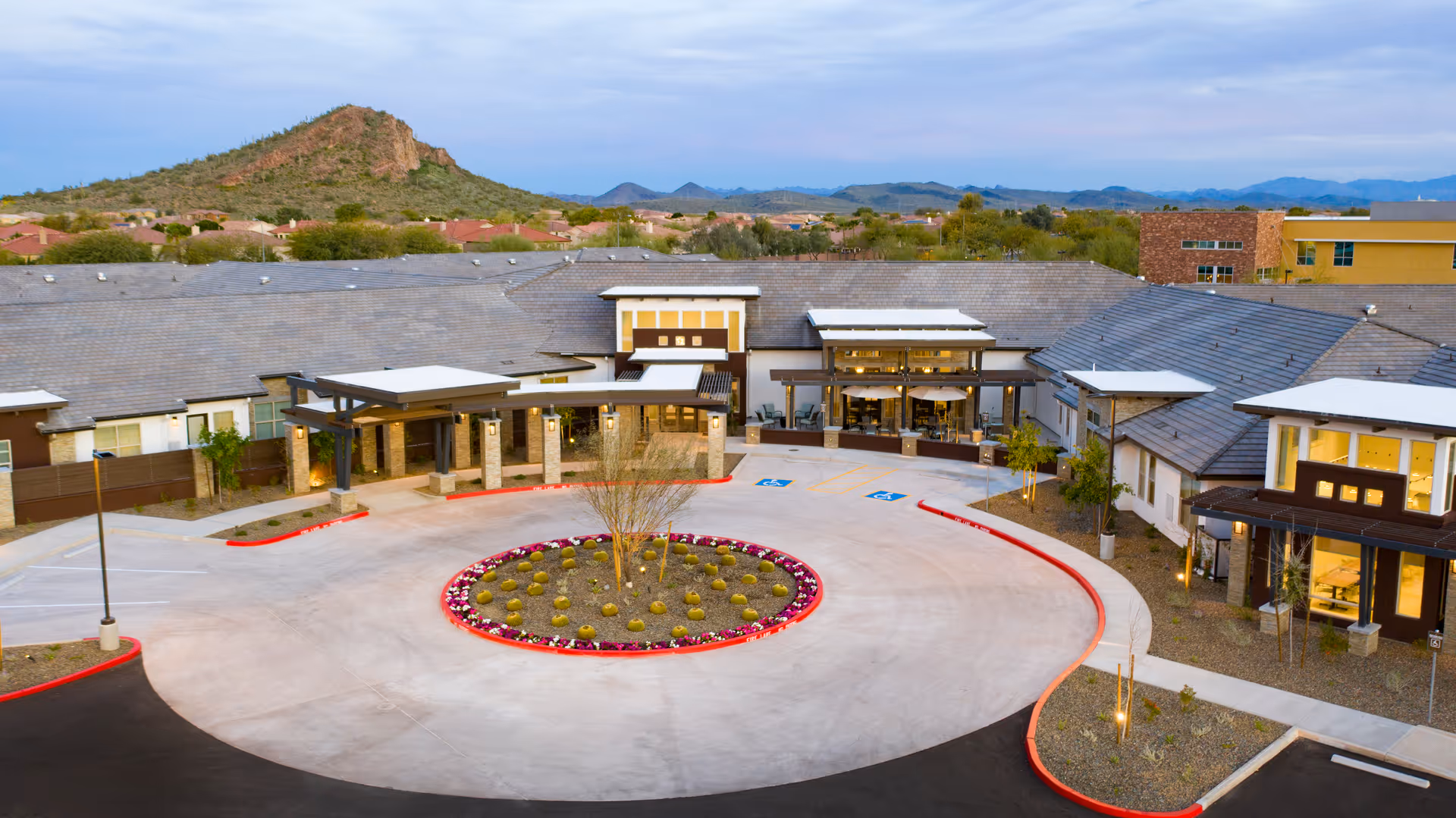 Aerial view of a senior living facility's front entrance with a circular driveway, landscaped center island, and surrounding desert hills.