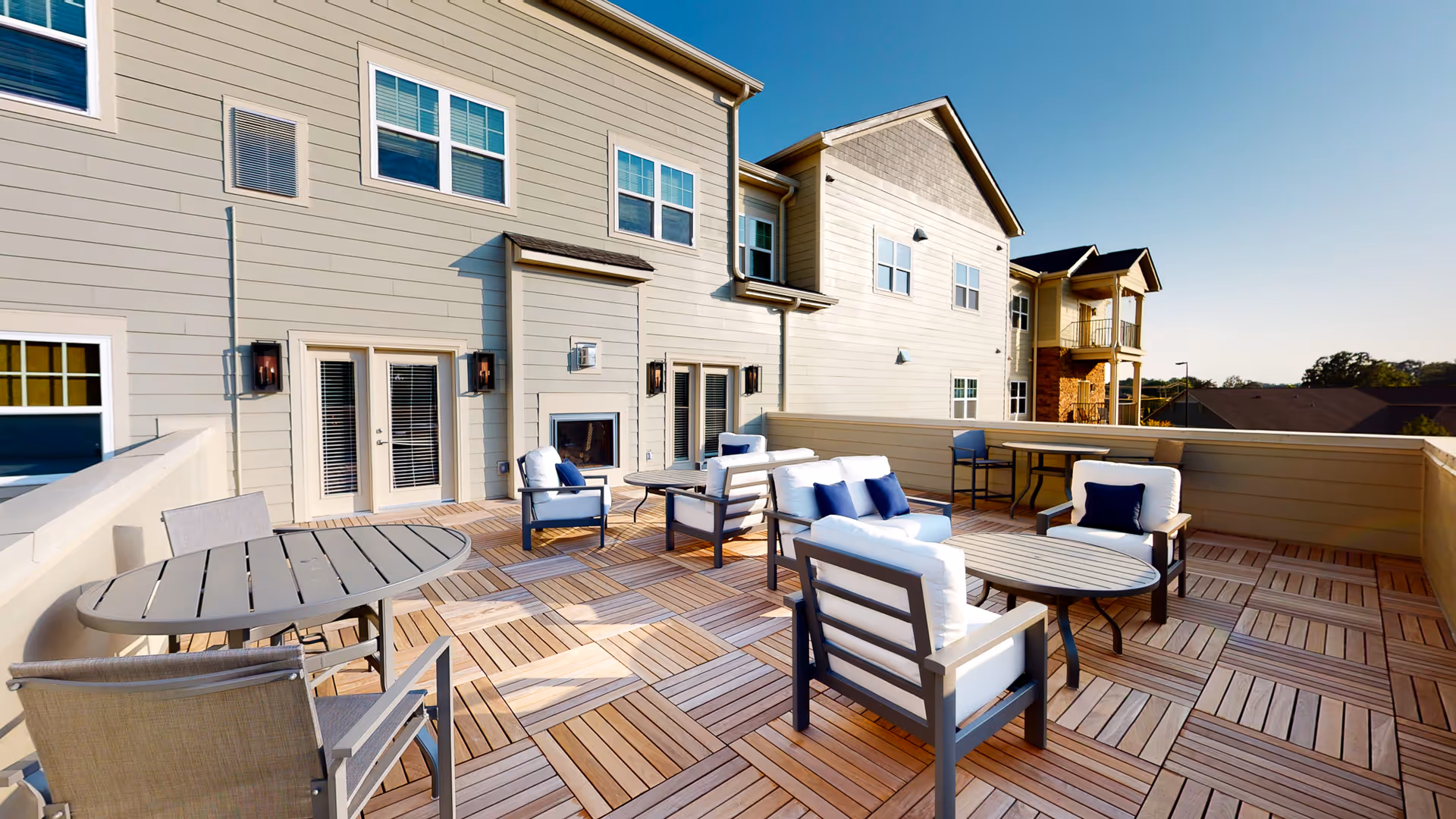 Spacious outdoor patio area with wooden tile flooring, several cushioned chairs with blue pillows, round tables, and a fireplace mounted on the exterior wall of a beige building under a clear blue sky.