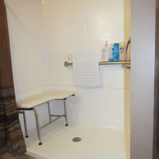 A clean shower area with a white bench seat, a grab bar with a white towel hanging on it, and several bottles of toiletries placed on the grab bar.