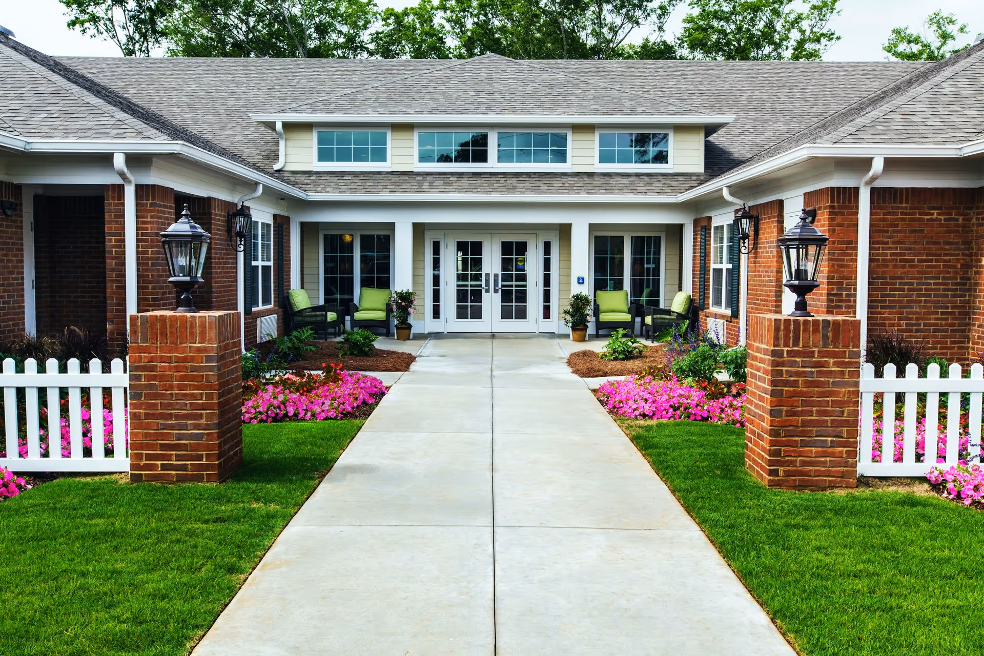 Front entrance of a brick memory care residence with a central walkway, white double doors, porch seating, brick pillars and colorful flowerbeds.