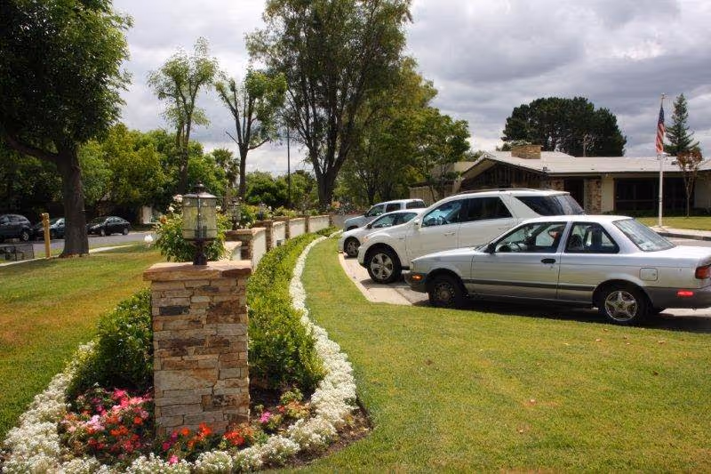 Landscaped front exterior with stone pillars, flowerbeds, parked cars, and a low building under a cloudy sky.