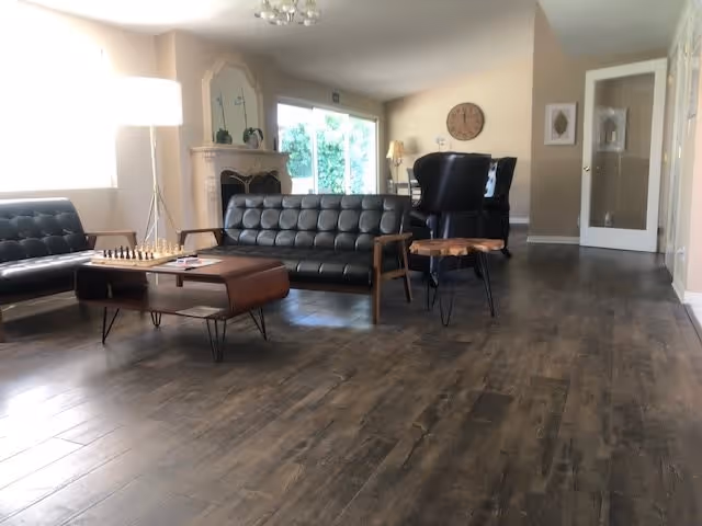 A spacious living room with dark wooden flooring, featuring two black leather tufted sofas, a wooden coffee table with a chessboard on it, a small wooden side table, a fireplace with decorative items on the mantel, a floor lamp, and a large window letting in natural light.