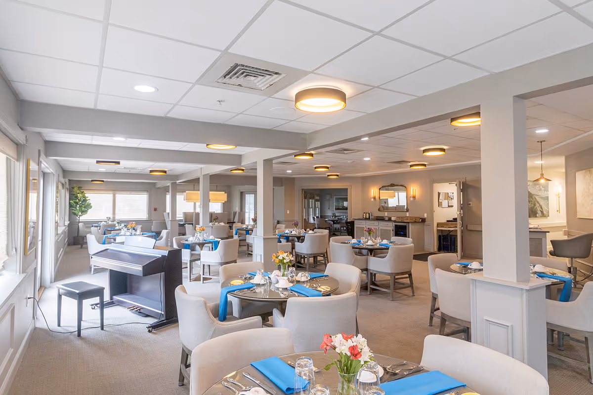 Spacious senior living dining room with round tables set with blue napkins, upholstered chairs, and a piano by the windows.