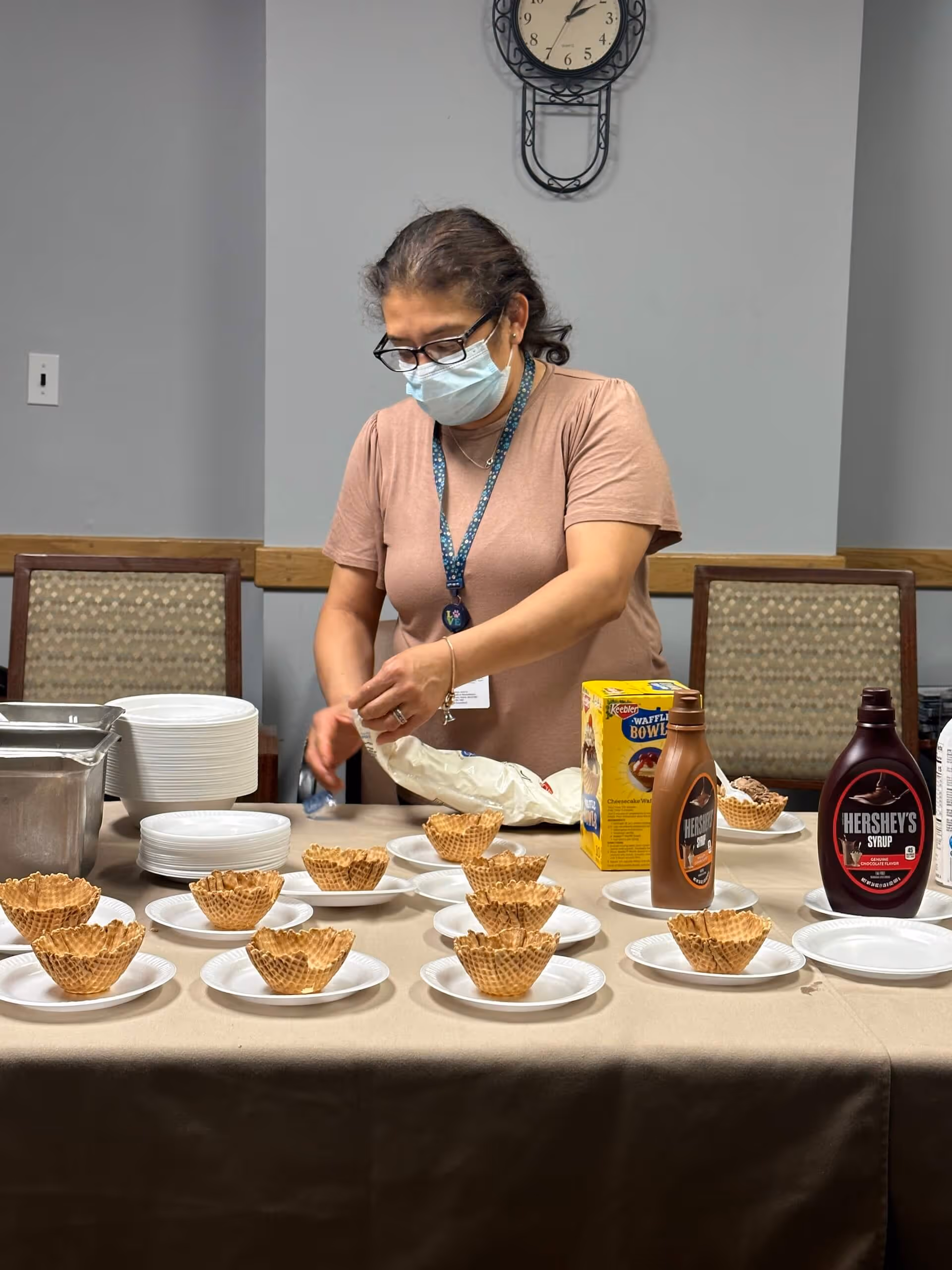 A woman wearing a face mask and glasses is preparing waffle bowls on a table. The table has several waffle bowls placed on white plates, along with bottles of Hershey's syrup and a box of Keebler Waffle Bowls. The setting appears to be an indoor room with chairs and a clock on the wall in the background.