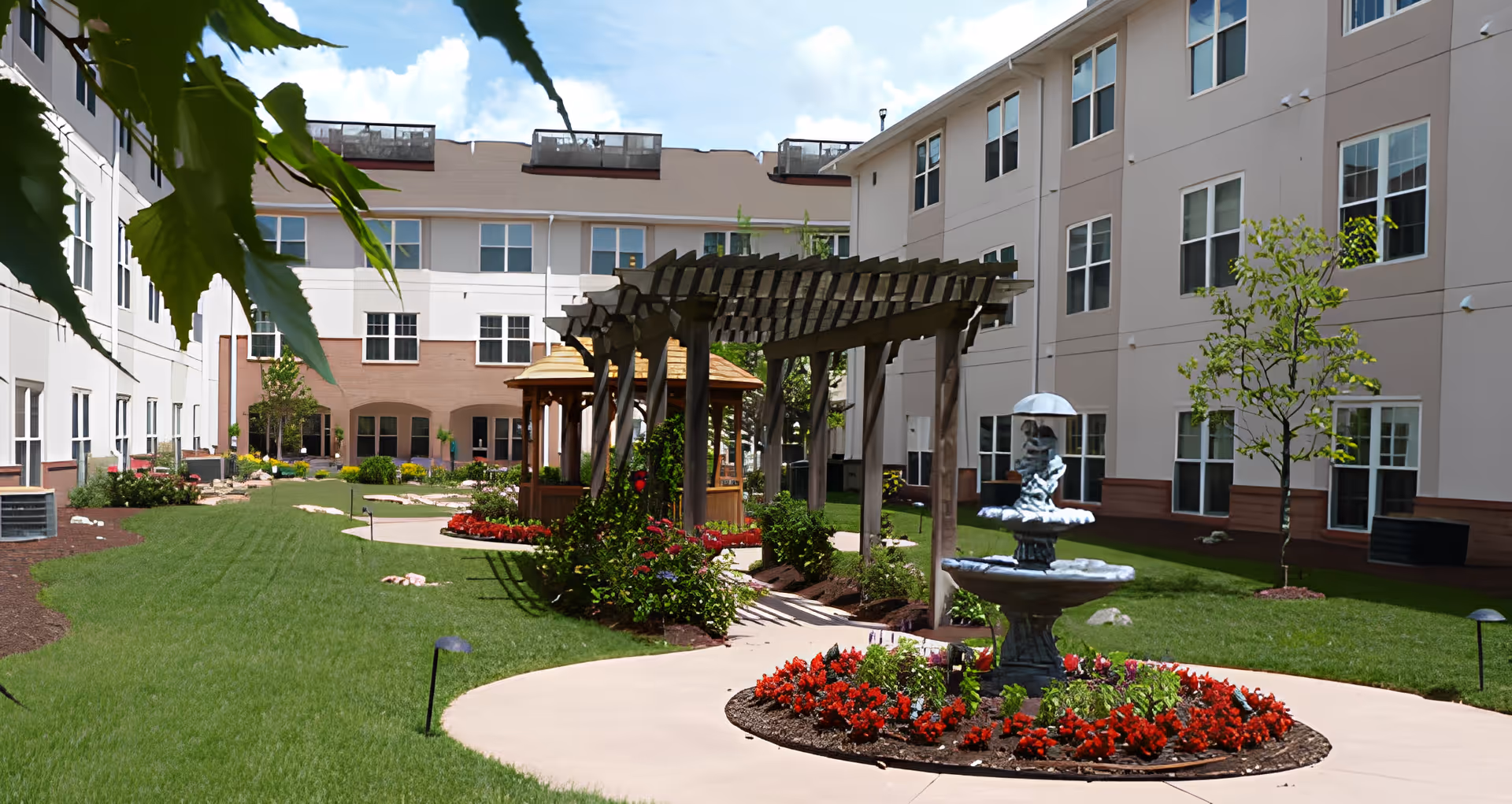 Landscaped courtyard of a multi-story retirement community with a pergola, fountain, flowerbeds and walkways between buildings.