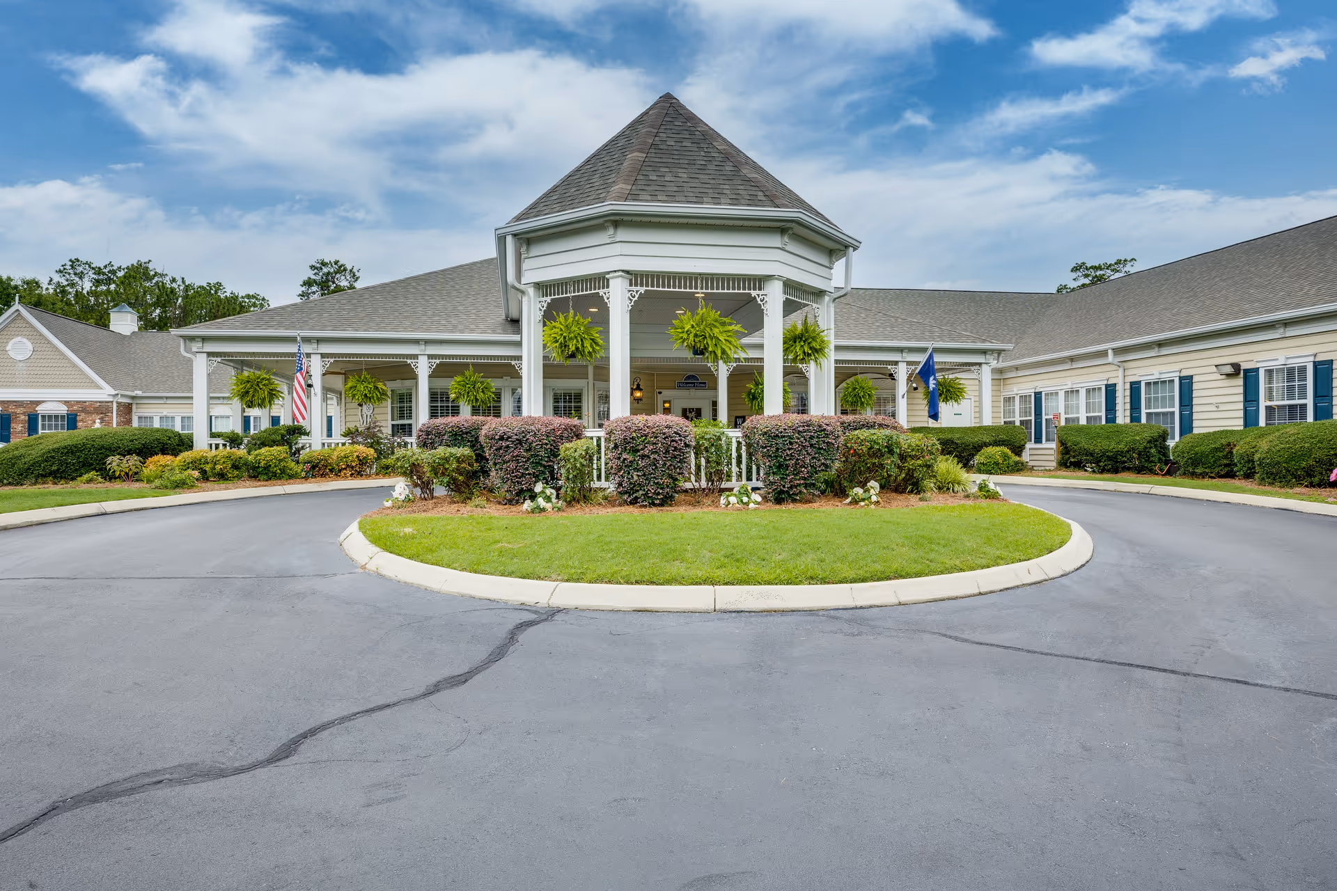 Front exterior view of The Legacy of Camden facility showing a circular driveway with a landscaped island in the center, a covered entrance with hanging plants, and a building with beige siding and blue shutters under a partly cloudy sky.