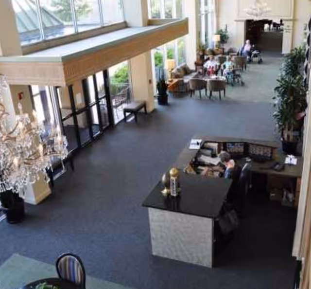 Interior view of a spacious senior living facility lobby with a reception desk where a staff member is seated. The area features large windows allowing natural light, a chandelier, seating areas with chairs and tables, and plants along the corridor.