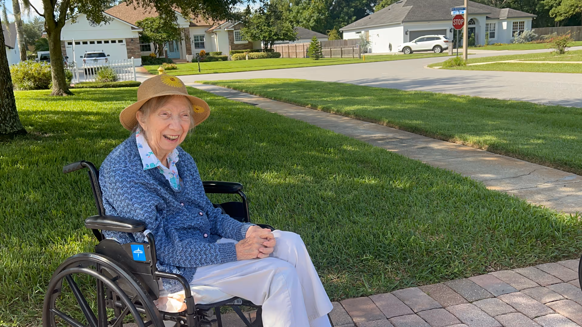 An elderly woman sitting in a wheelchair on a paved pathway in a residential neighborhood. She is wearing a blue cardigan, white pants, and a straw hat with a yellow flower, smiling and enjoying the sunny day. Green grass, trees, and houses are visible in the background.
