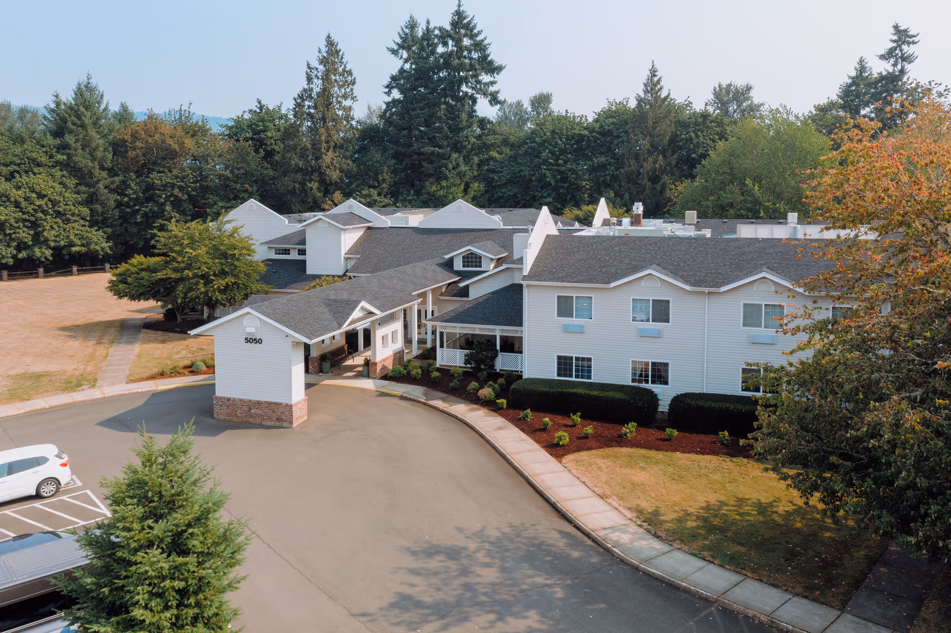 Aerial view of Wiley Creek Senior Living facility showing a large white multi-story building with a covered entrance, surrounded by trees and landscaped bushes. There is a parking area with a few cars and a driveway leading to the entrance.