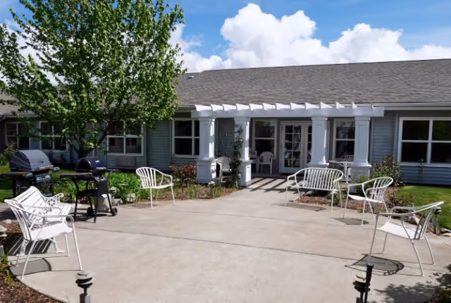 Outdoor courtyard patio with white chairs, grills, and a pergola in front of a single-story senior living building.
