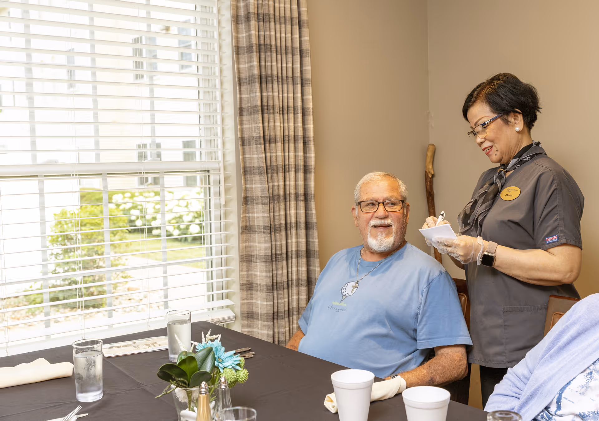 An elderly man wearing glasses and a blue shirt sits at a dining table with a black tablecloth, looking up and smiling at a female staff member who is standing next to him taking notes. The room has a large window with white blinds and plaid curtains, and there are glasses of water and white cups on the table.