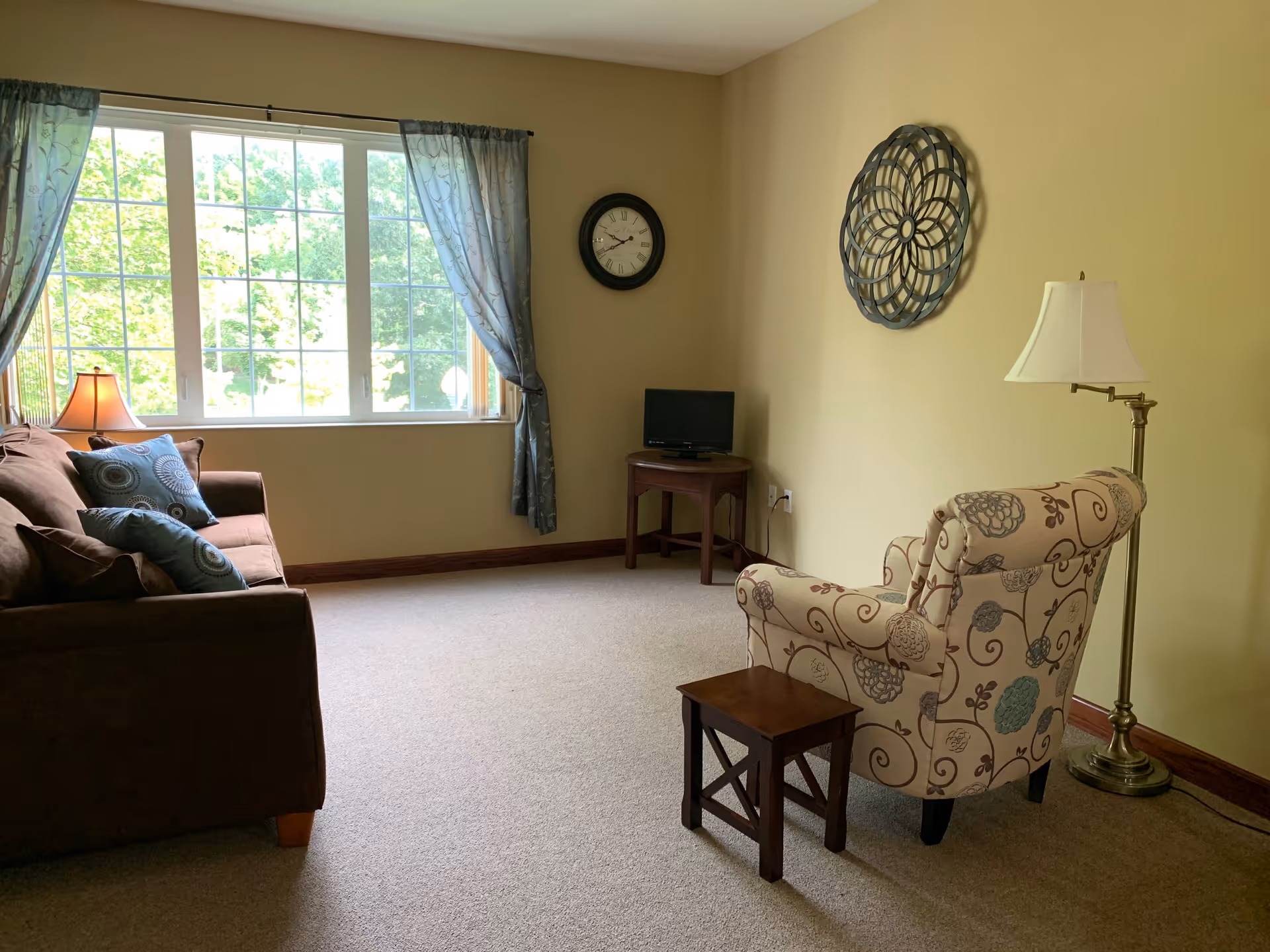 A cozy living room with a large window covered by sheer curtains letting in natural light. The room features a brown sofa with patterned cushions, a floral-patterned armchair, a small wooden side table, a floor lamp, a wall clock, and a decorative wall piece. A small TV sits on a wooden corner table.