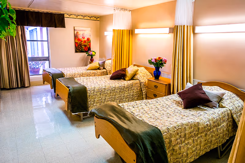 A bright and clean room with three single beds arranged in a row, each with patterned bedspreads, pillows, and dark green throws at the foot. Between the beds are wooden nightstands with vases of colorful flowers. The room has large windows with curtains and soft wall lighting, creating a warm and welcoming atmosphere.