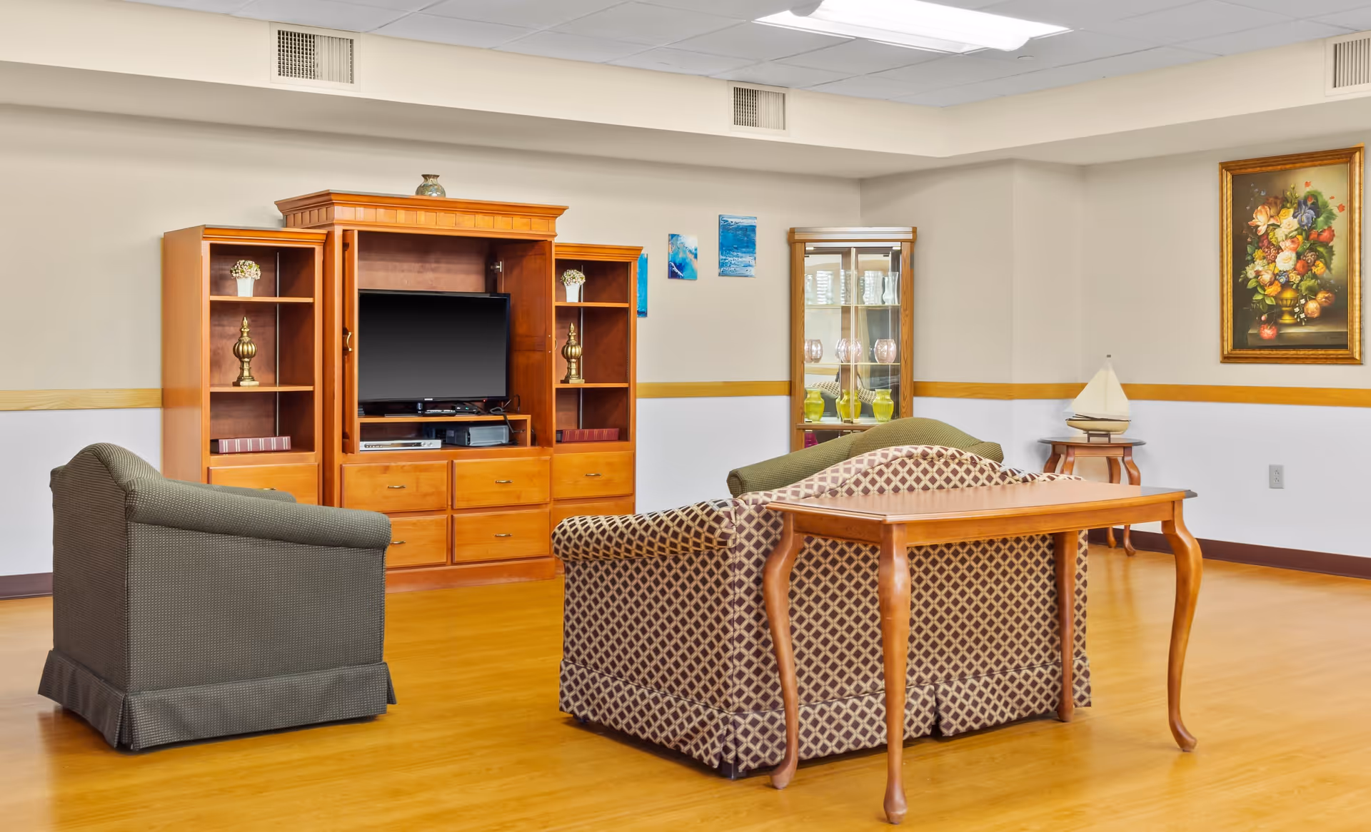 A senior living facility common area with two upholstered armchairs and a patterned loveseat arranged around a wooden entertainment center with a flat-screen TV. The room has light-colored walls with a wooden chair rail, a glass display cabinet with decorative glassware, a small side table with a model sailboat, and a framed floral painting on the wall.