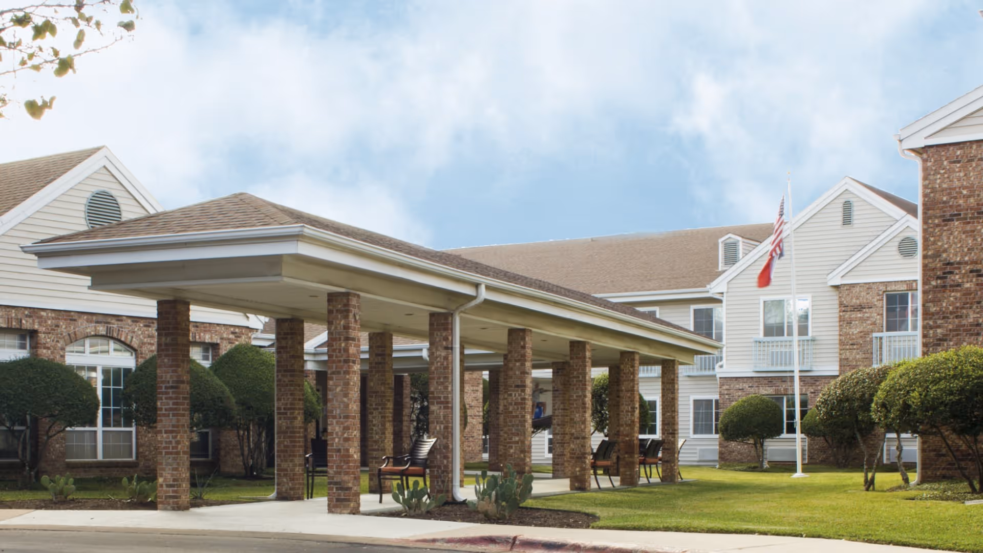 Exterior view of Juniper Village at Spicewood Summit showing a covered entrance supported by brick columns, manicured bushes, green lawn, and a flagpole with the American flag in front of a two-story building with brick and white siding.