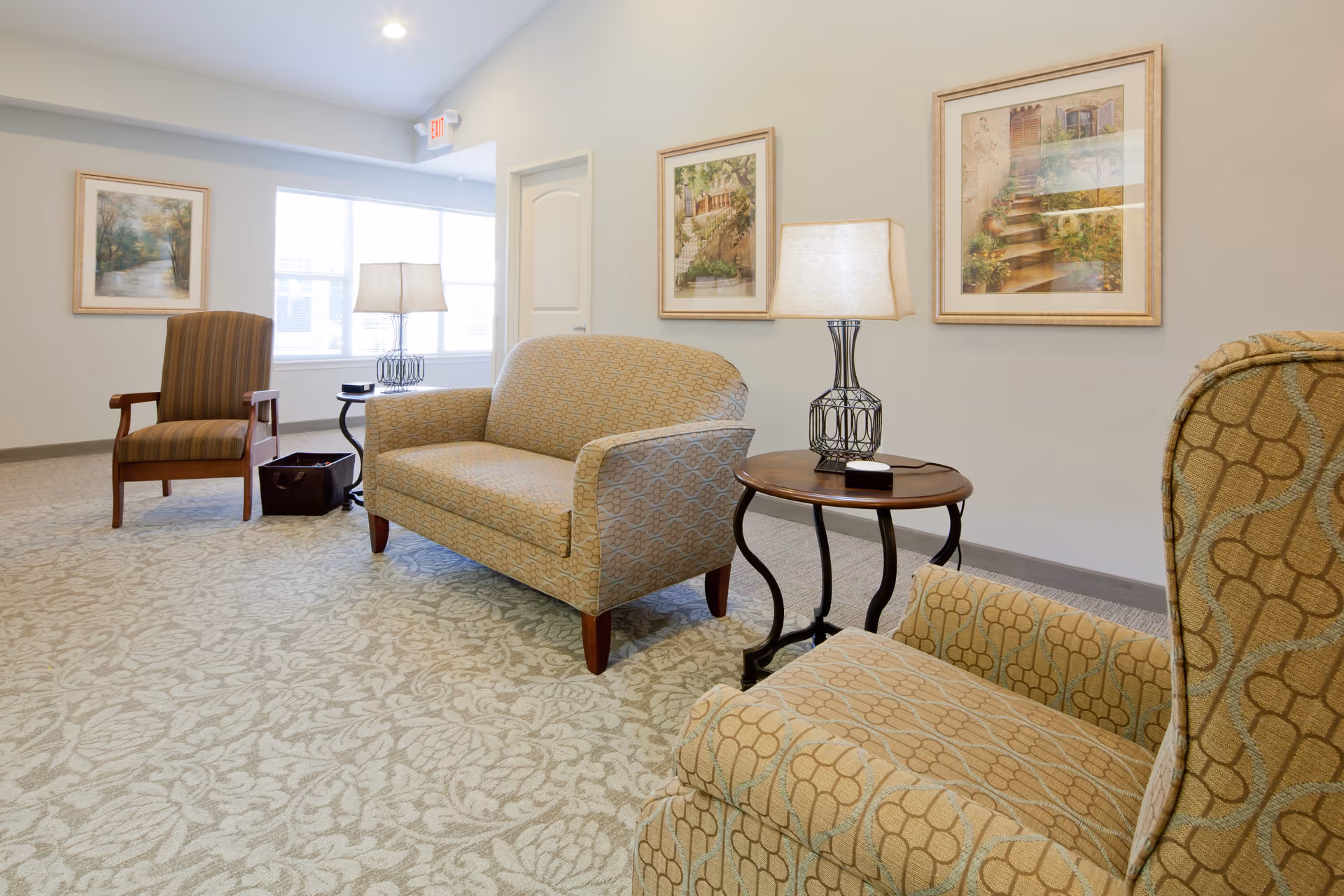 A cozy sitting area in a senior living facility with patterned armchairs and a loveseat arranged around a small wooden side table with a lamp. The room has light-colored walls adorned with framed artwork and a large window letting in natural light.