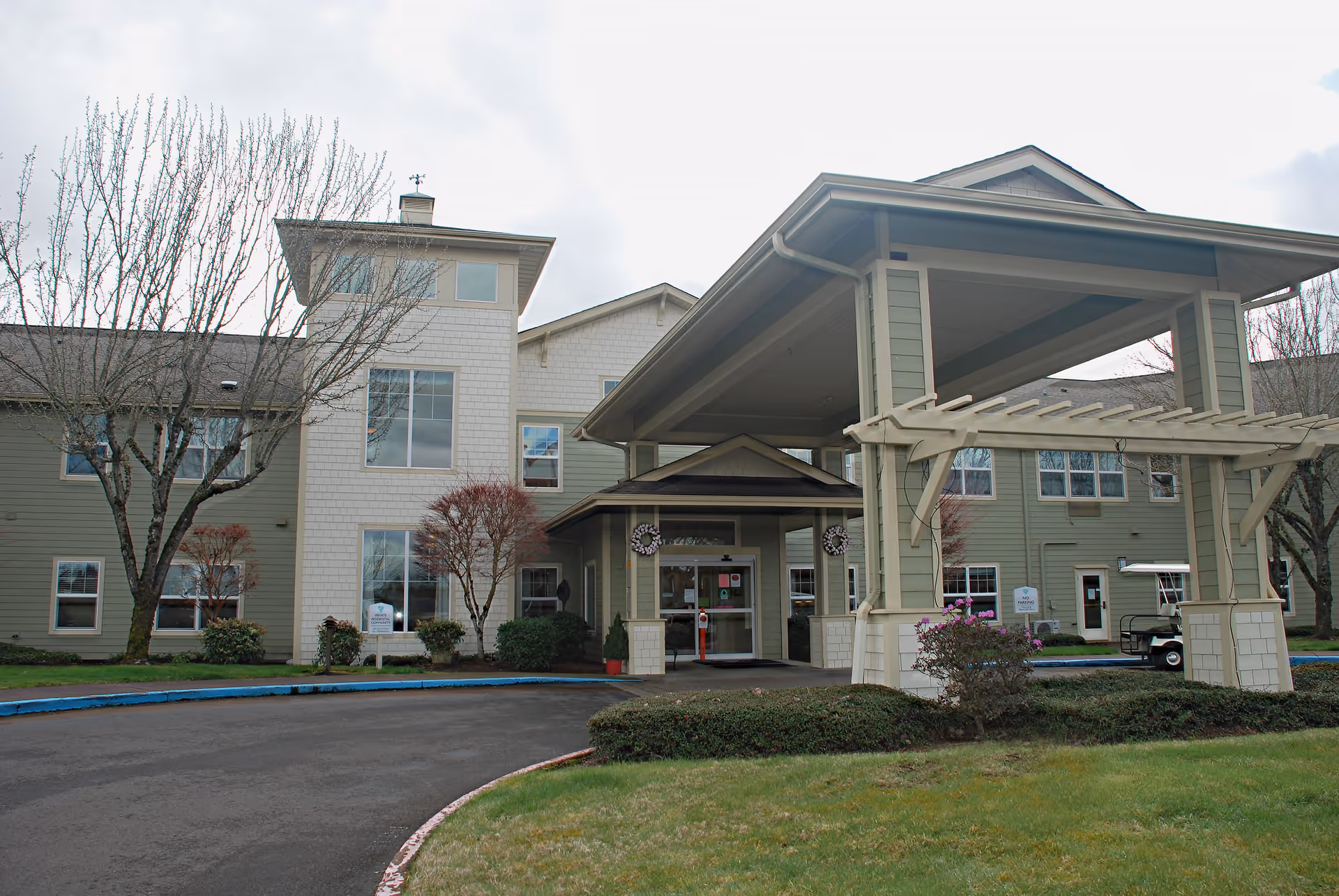 Exterior view of Cascades of Stayton senior living facility showing the main entrance with a covered drop-off area, surrounded by trees and shrubs, under a cloudy sky.