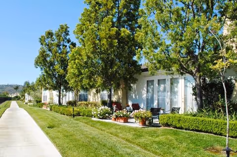 Sunlit row of single-story residential units with patios, potted plants, and a manicured lawn and walkway.