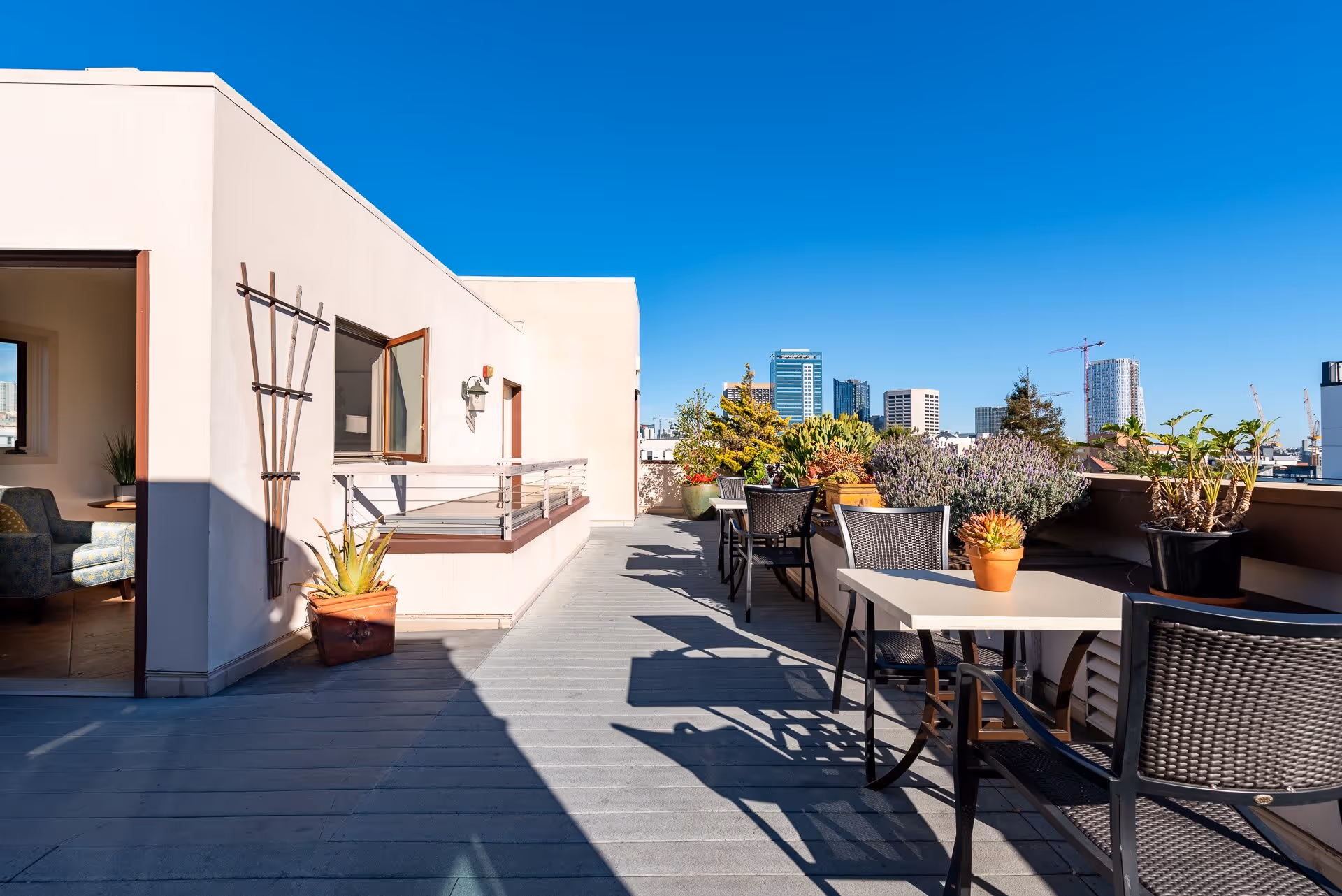 Outdoor patio area at The Village at Hayes Valley with tables and chairs, potted plants along the railing, and a clear view of city buildings under a bright blue sky.