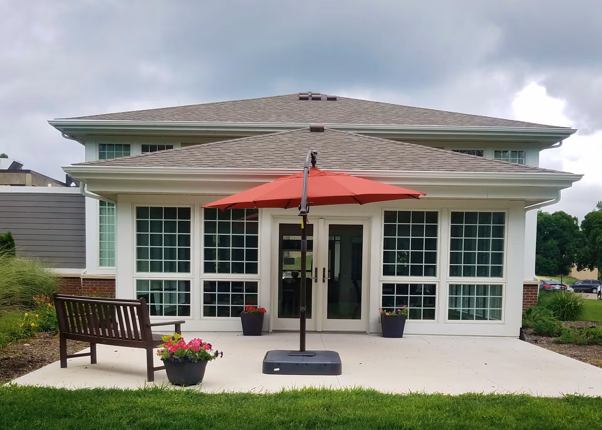 Exterior view of a building with large windows and double glass doors. In front of the building is a concrete patio with a red umbrella, a wooden bench, and flower pots with colorful flowers. The sky is cloudy and there is green grass and landscaping around the patio.