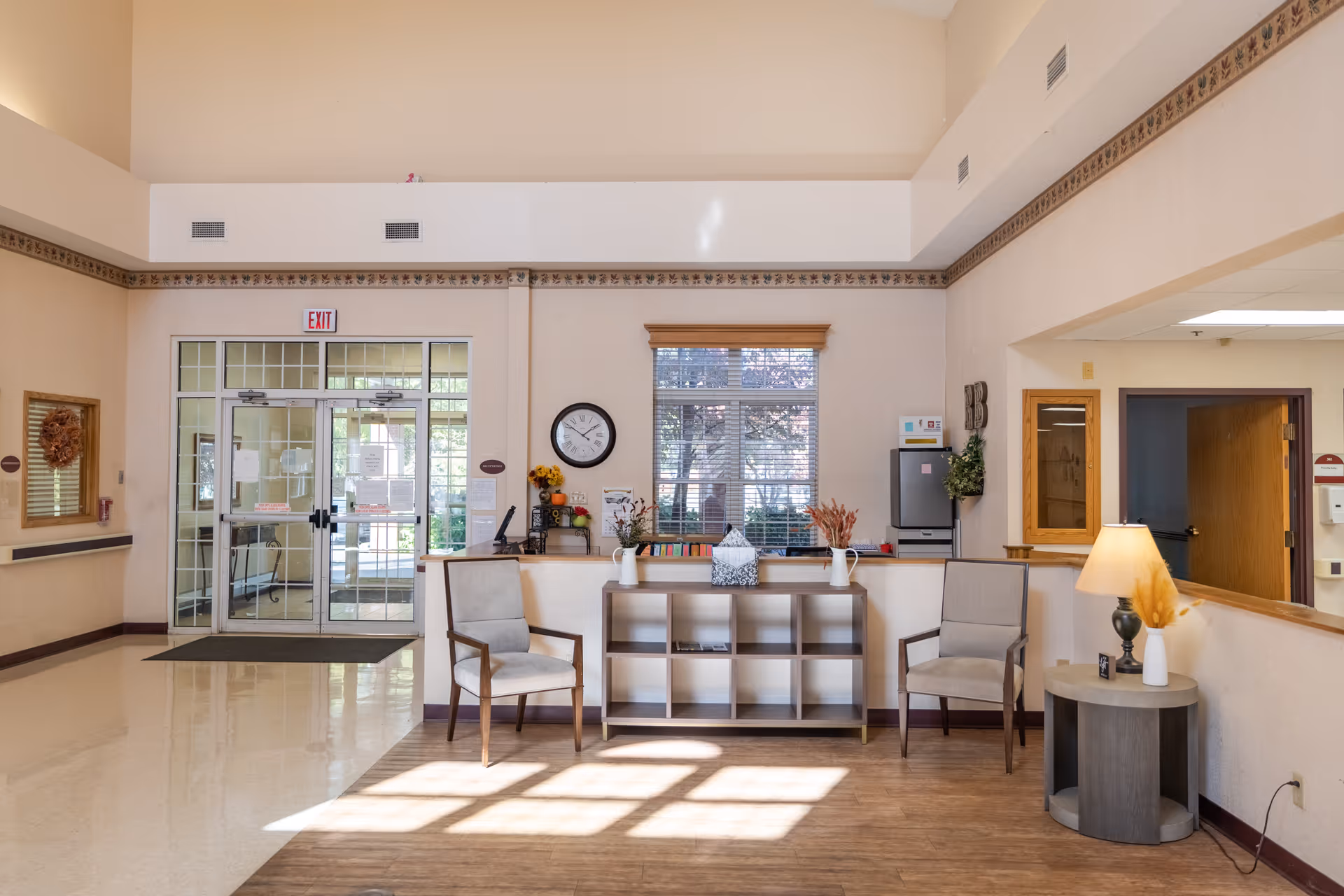 A bright and clean nursing facility lobby area with two cushioned chairs on either side of a small wooden shelf unit. The shelf holds decorative items including a tissue box and vases with dried flowers. A round side table with a lamp and a small decorative item is next to one chair. Behind the shelf is a window with blinds, a wall clock, and a small refrigerator. Double glass doors with an exit sign above lead outside. The walls are light beige with a decorative border near the ceiling.