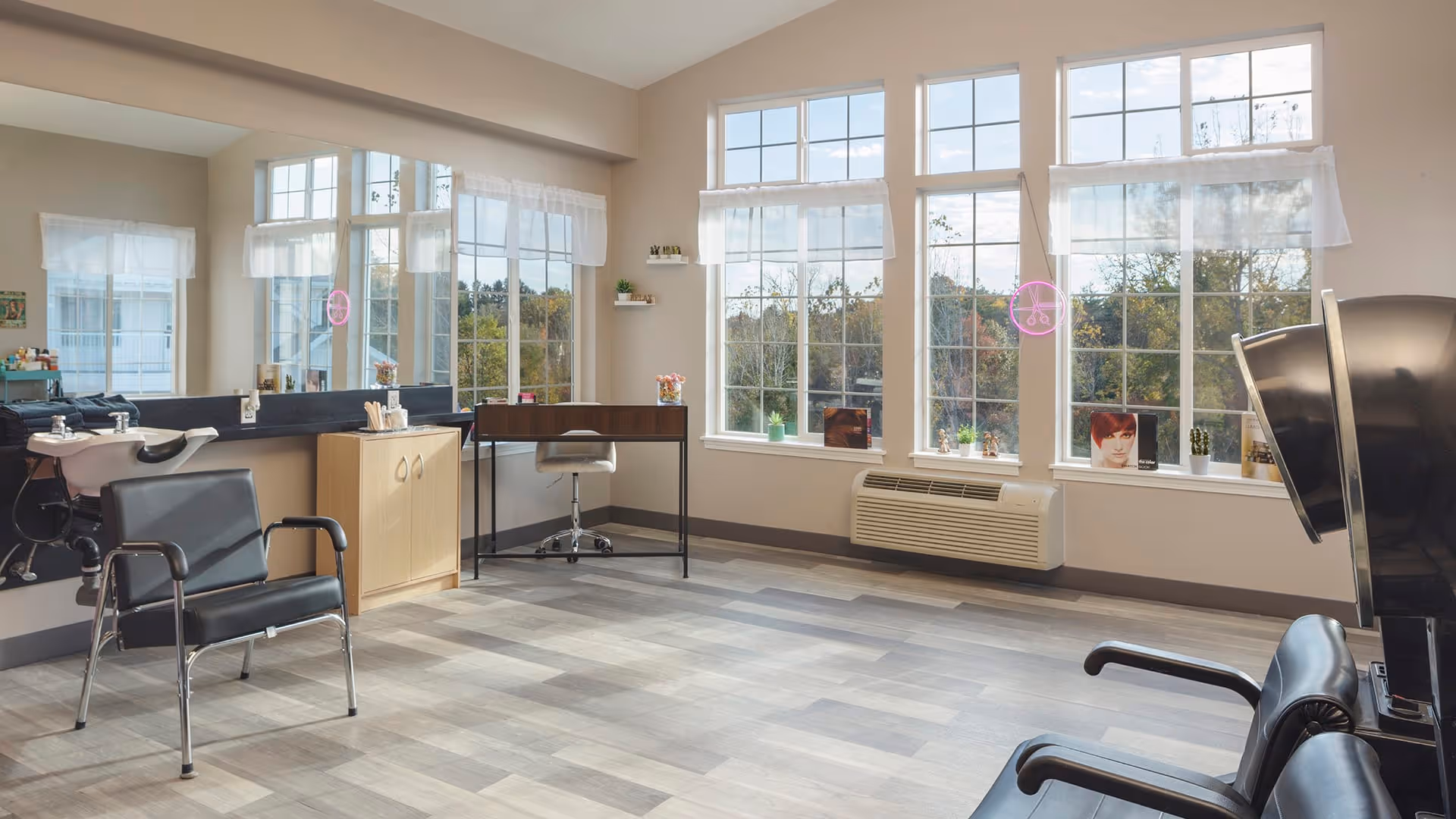 A bright and spacious hair salon area with large windows letting in natural light. The room features black salon chairs, a hair washing station, a small desk with a chair, and various hair care products on the counter. The windows have sheer white curtains and small potted plants on the windowsills.