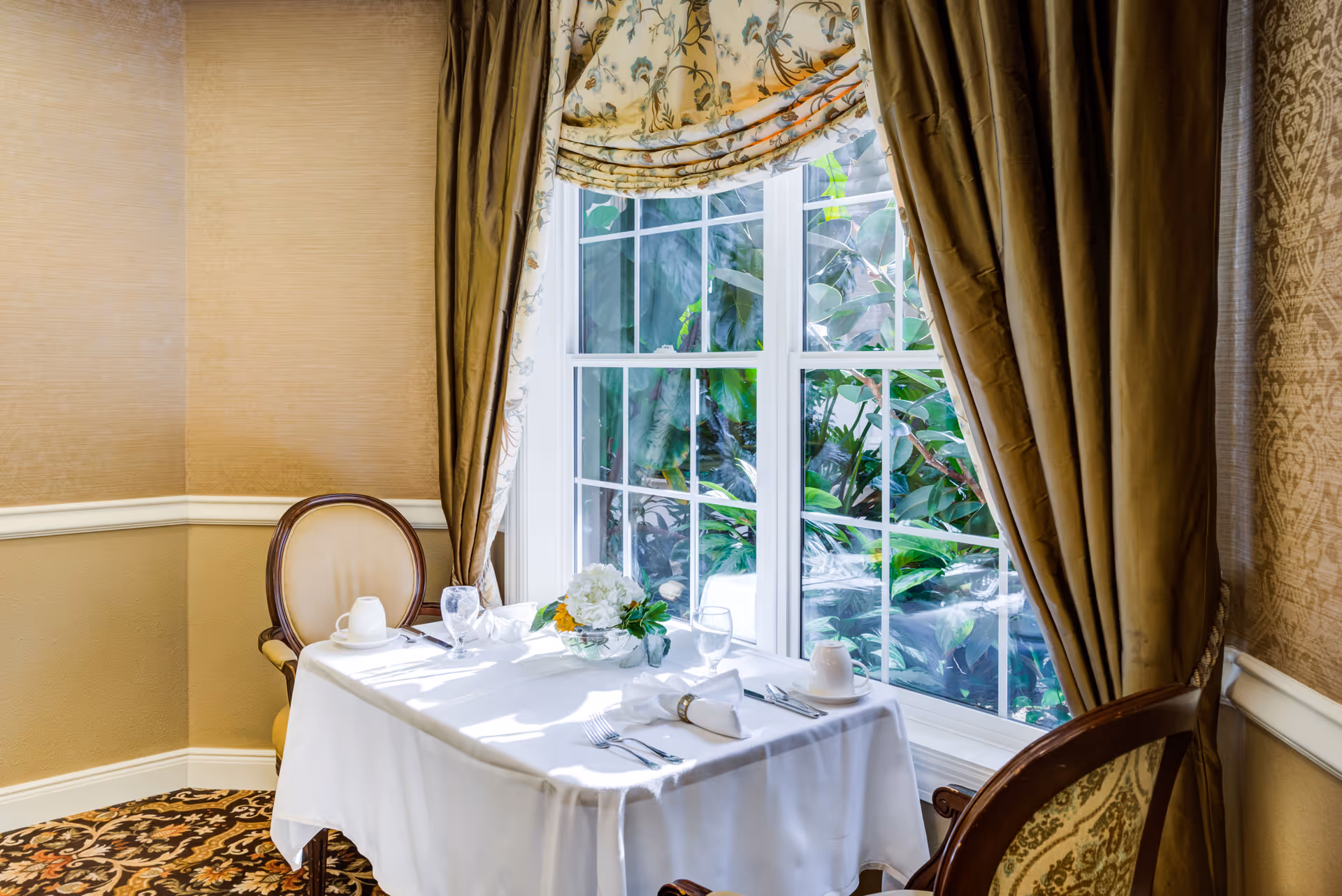 A small dining table set for two with white tablecloth, napkins, cups, glasses, and silverware, placed next to a large window with floral curtains. Outside the window, green plants are visible. The room has beige walls and patterned carpet.