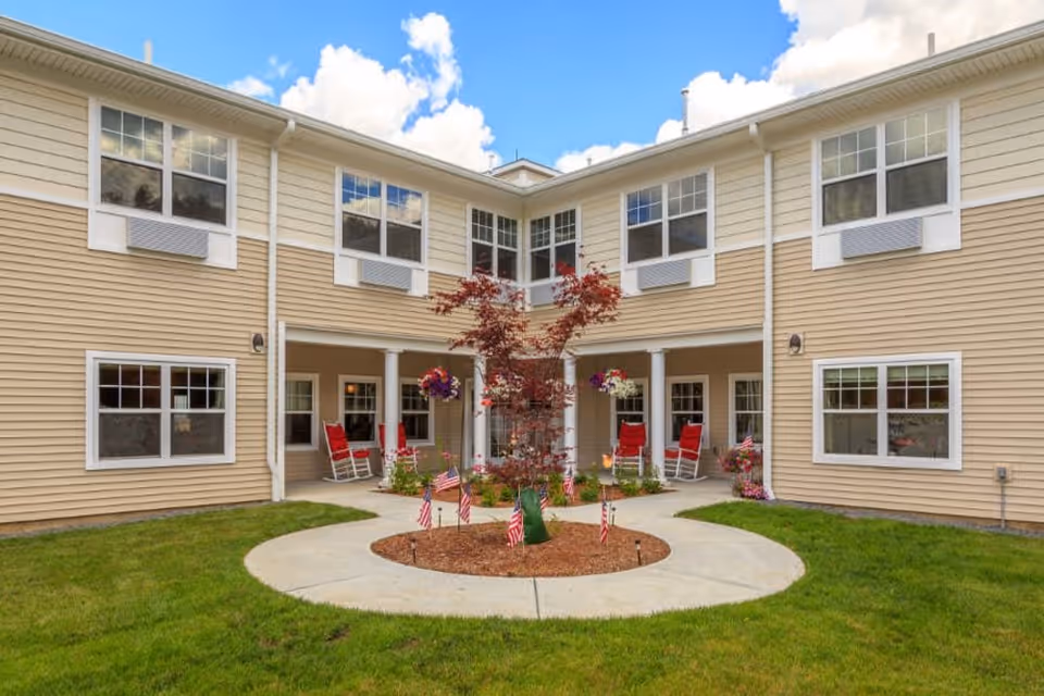 Outdoor courtyard area of a senior living facility with a circular concrete pathway surrounding a small tree decorated with miniature American flags. The building is two stories with beige siding and white trim. There are red rocking chairs and hanging flower baskets on the covered porch area.