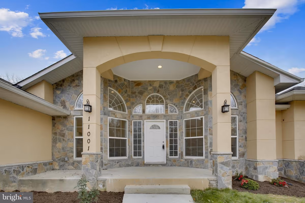 Front exterior view of a senior care facility entrance with a covered porch supported by stone and beige pillars, a white door with multiple windows surrounding it, and a well-maintained garden area.
