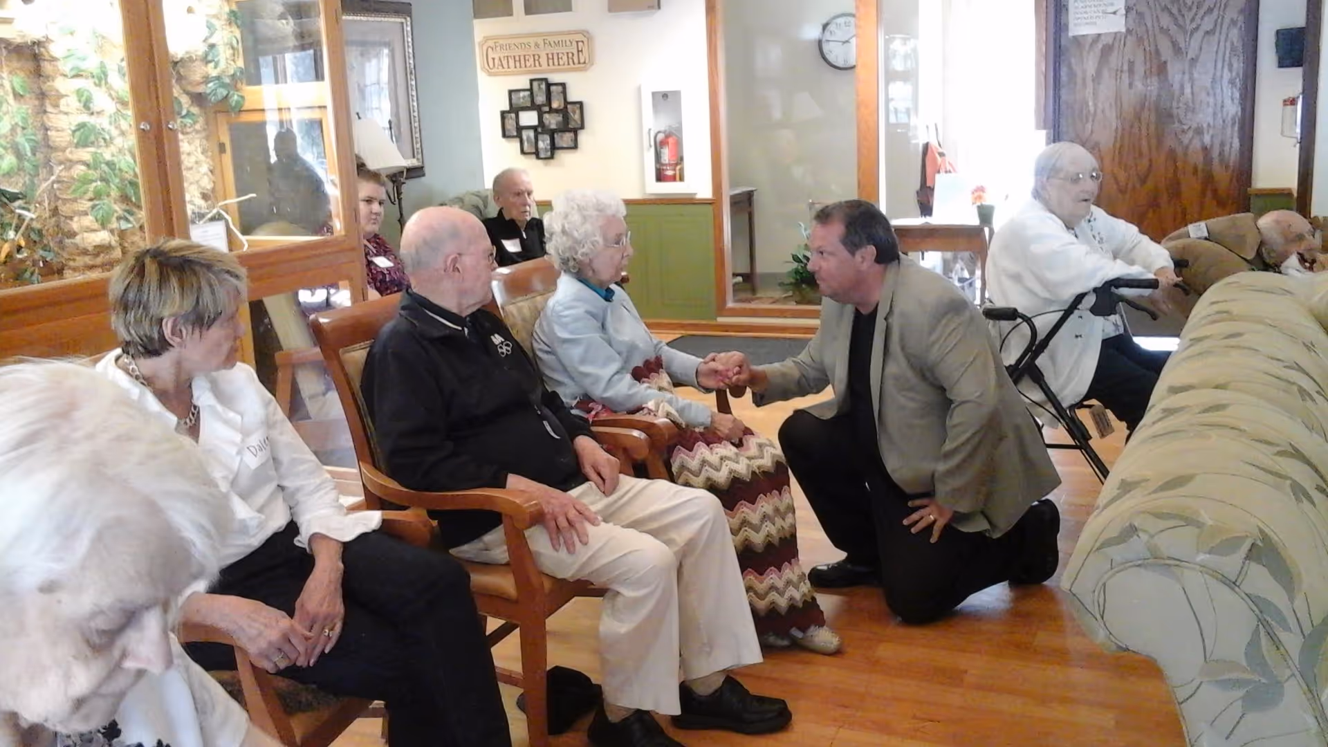 A group of elderly people sitting in chairs in a living room area, with one man kneeling and holding hands with an elderly woman. The room has wooden floors, a couch, and a sign on the wall that reads 'Friends & Family Gather Here'.