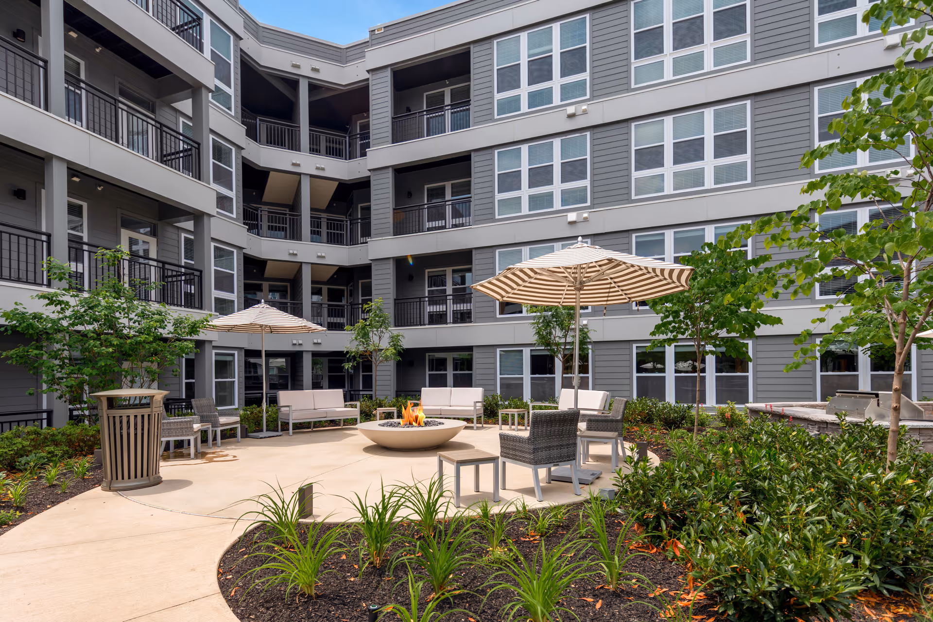 Courtyard patio with seating, umbrellas, and a central fire pit in front of a multi-story residential building.
