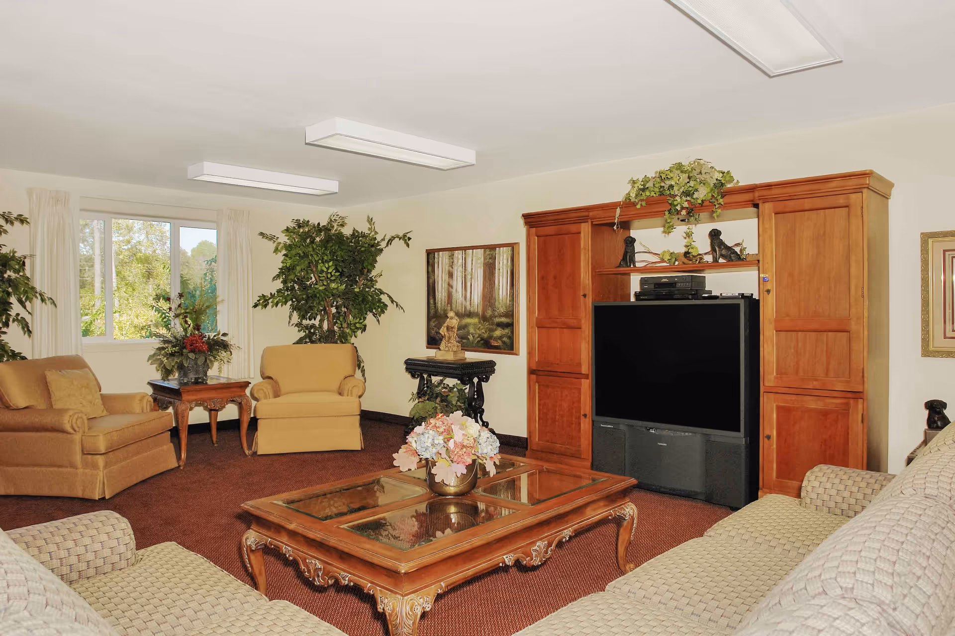 A cozy common living room with upholstered chairs and sofas arranged around a wooden coffee table and a large wooden entertainment center.