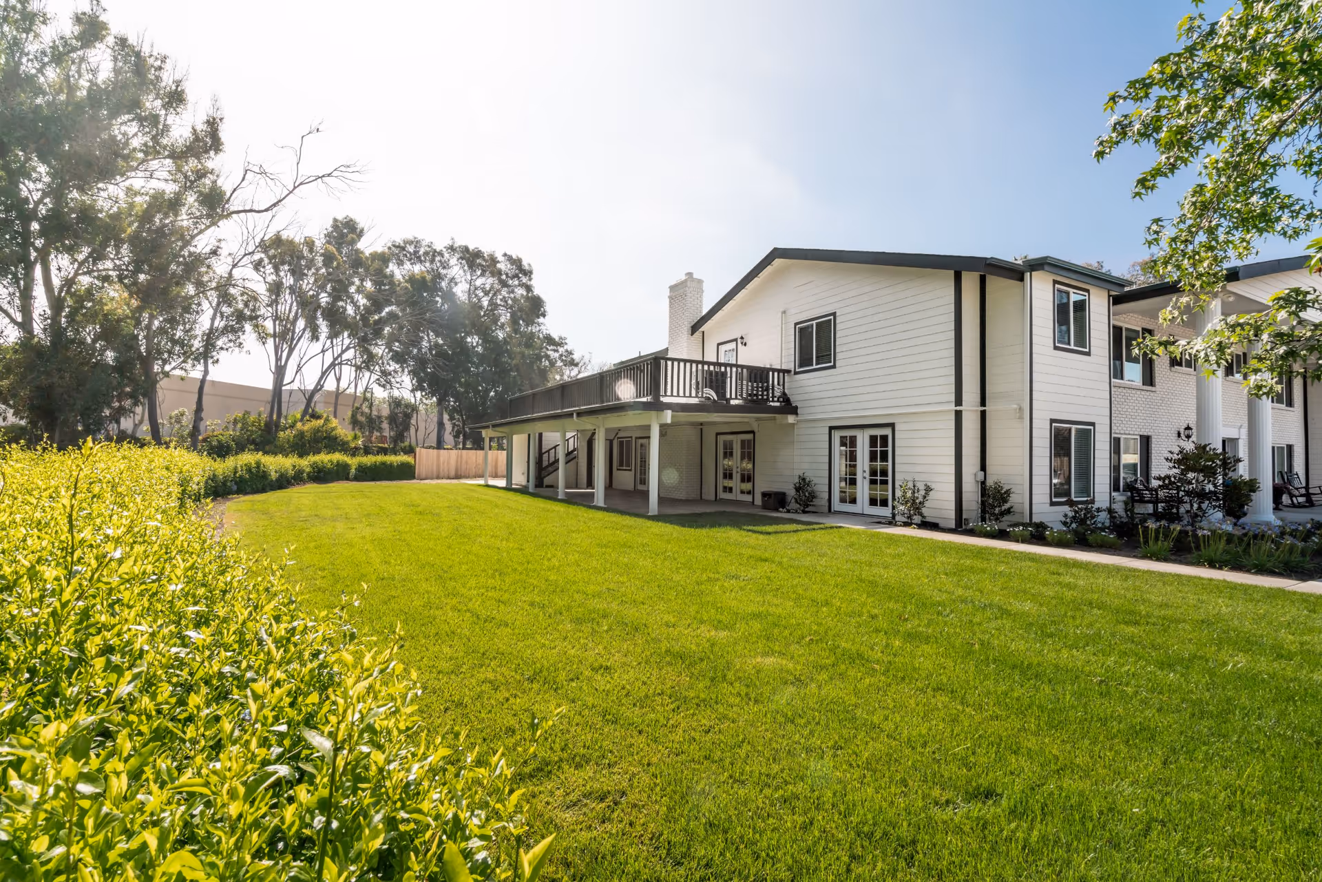 A bright outdoor view of a senior living facility building with white siding and multiple windows. The building has a covered patio area and an upper deck. The foreground features a well-maintained green lawn bordered by bushes and trees under a clear sky.