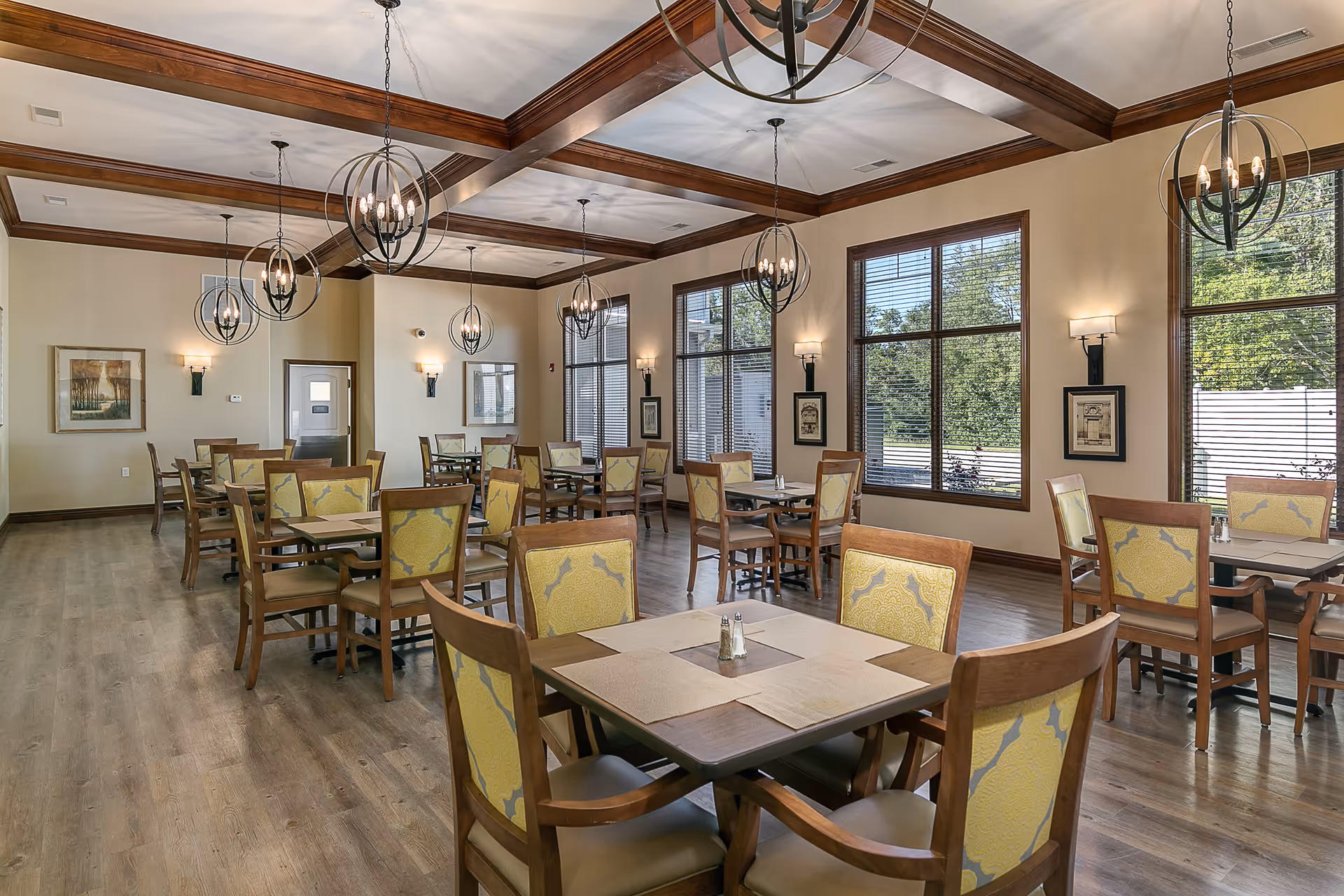 A spacious dining room with multiple wooden tables and chairs featuring yellow patterned upholstery. The room has large windows with blinds allowing natural light to enter, wooden ceiling beams, and several modern spherical chandeliers hanging from the ceiling. The walls are decorated with framed artwork and sconces.