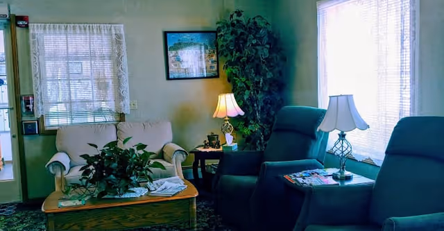A cozy living room area with two blue recliner chairs and a beige loveseat. There is a wooden coffee table with a plant and newspapers on it. Two table lamps are lit, one on a side table between the recliners and another on a small table next to the loveseat. A large potted plant is in the corner near a framed picture on the wall. The room has windows with sheer white curtains allowing natural light to enter.
