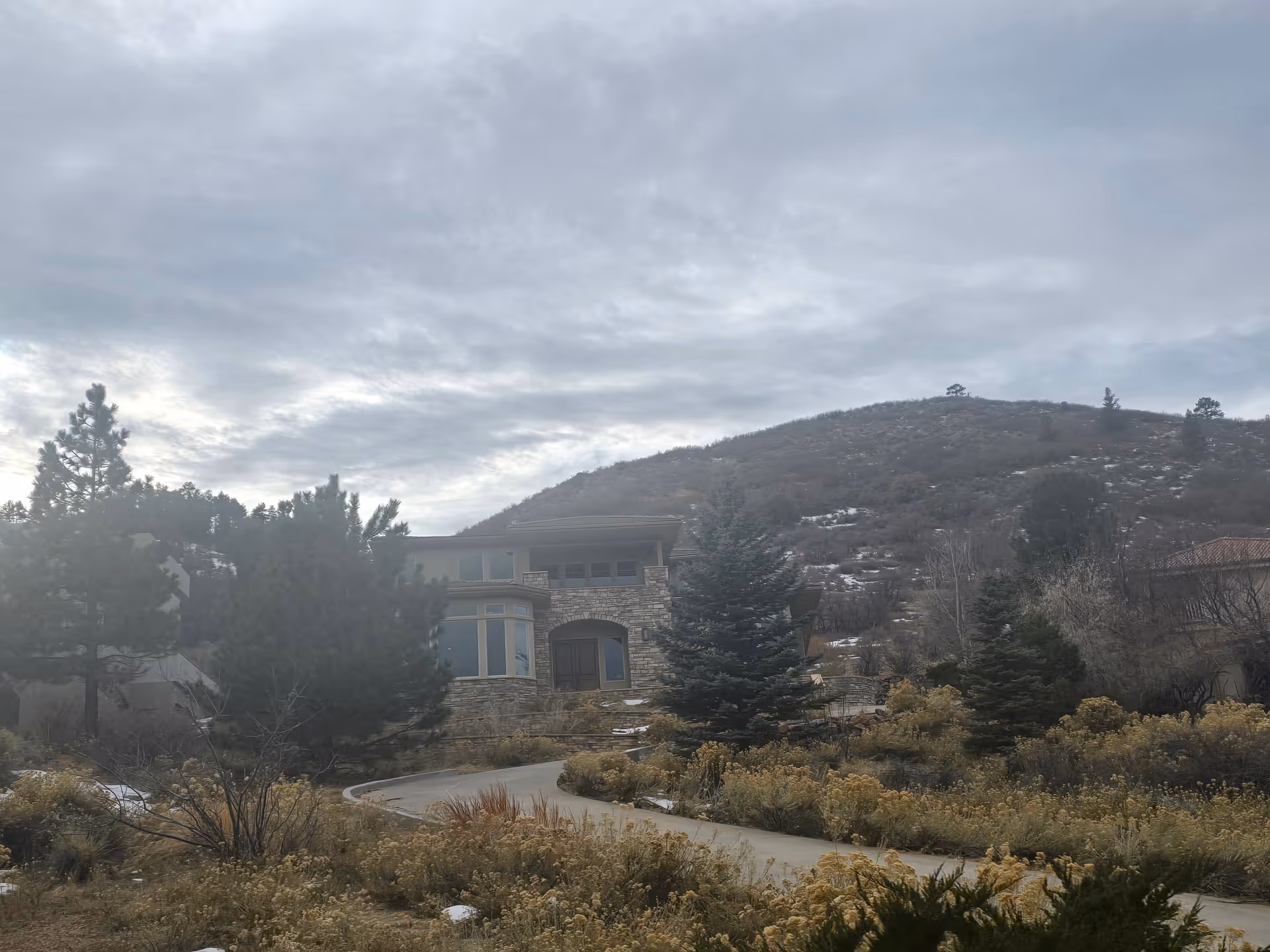 A large stone and wood house surrounded by trees and shrubs with a winding driveway in front. In the background, there is a hill with sparse vegetation under a cloudy sky.